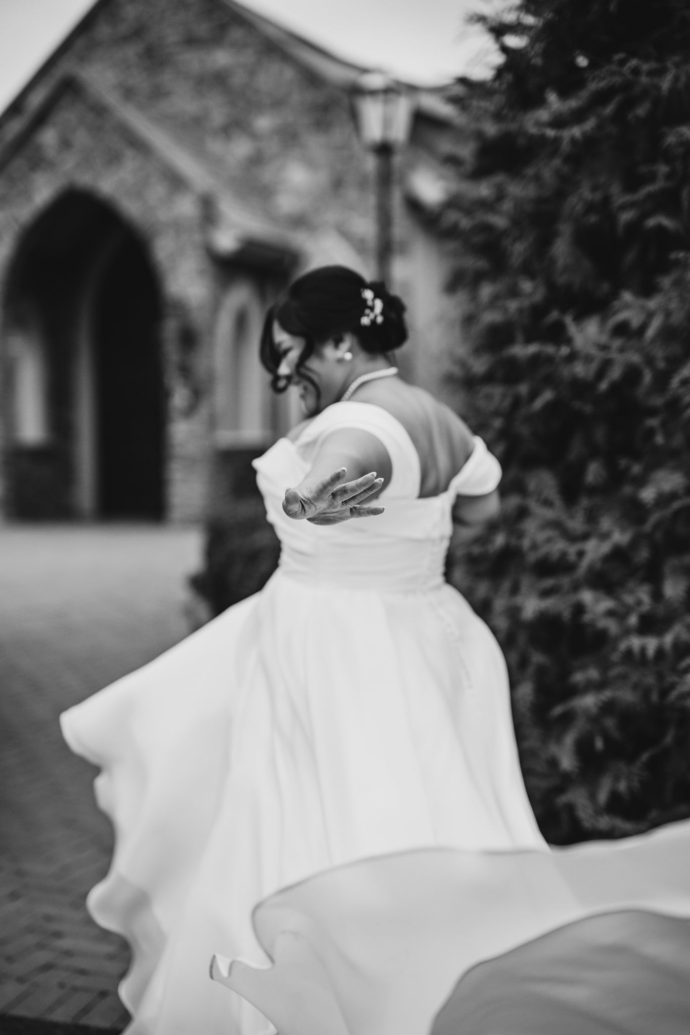 Black and white photo of bride twirling in flowing dress outside stone chapel.