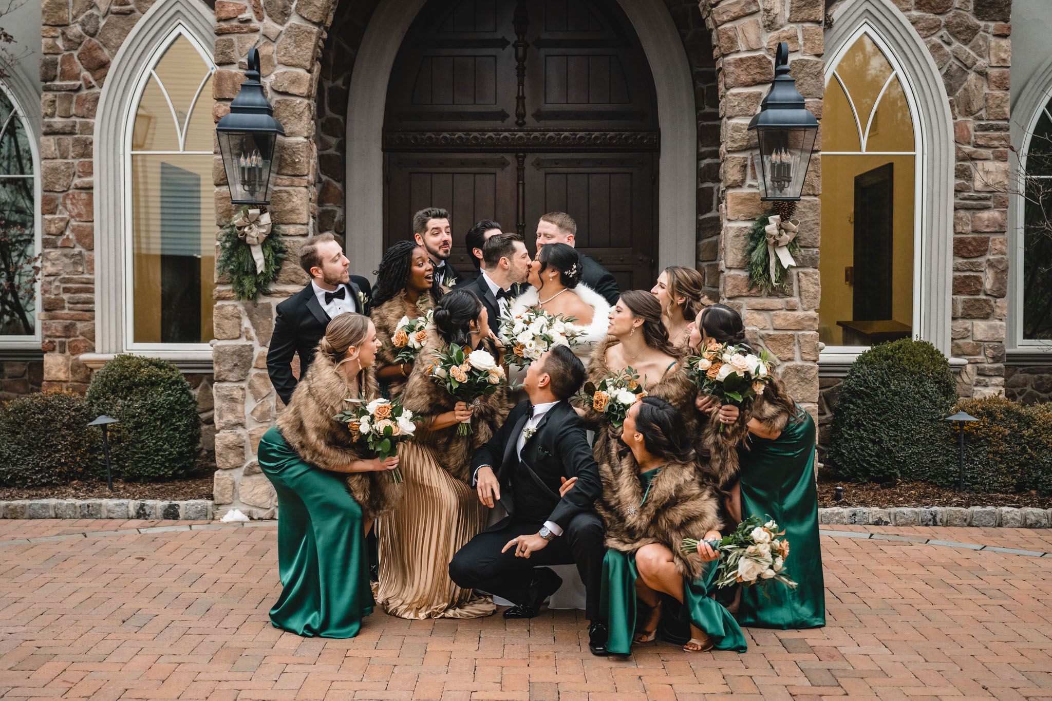 Wedding party poses joyfully in front of stone church with emerald green and gold attire.