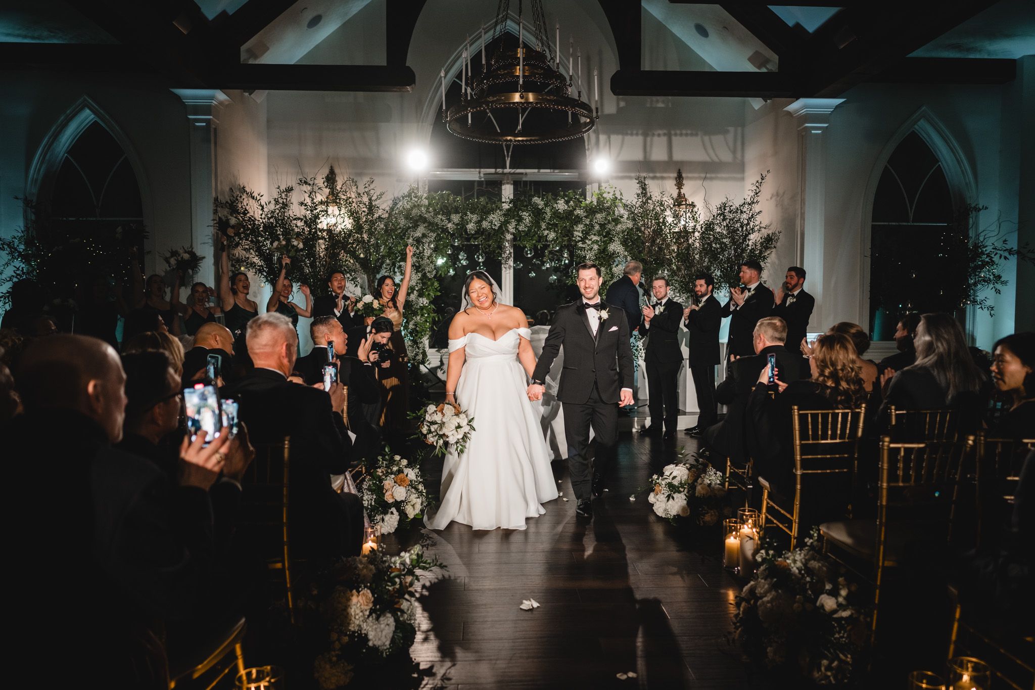 Newlyweds walk down aisle in modern church venue with dramatic lighting and floral decor.