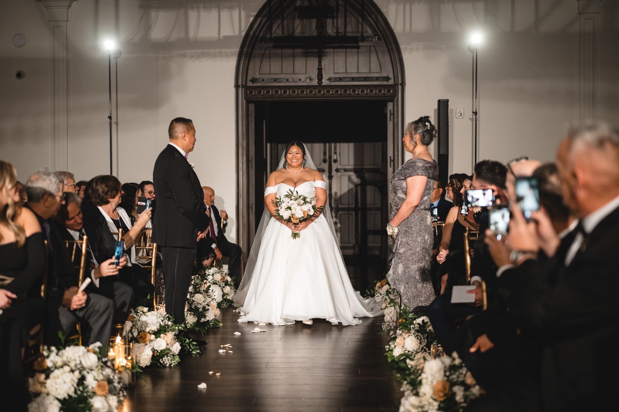 Candlelit wedding ceremony in a grand hall with guests capturing photos on phones.