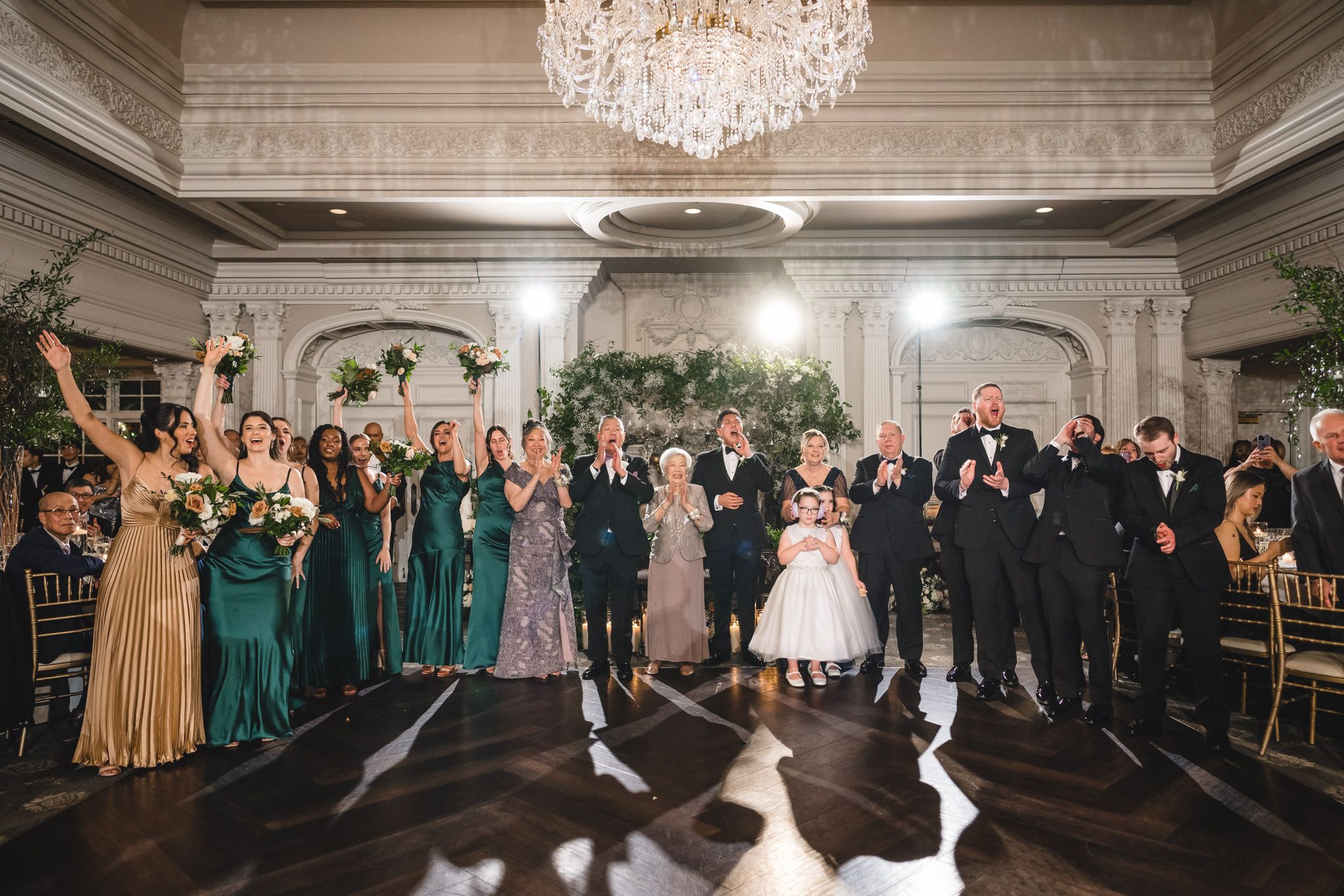 Wedding party celebrates in an elegant ballroom with crystal chandelier and dark hardwood floors.