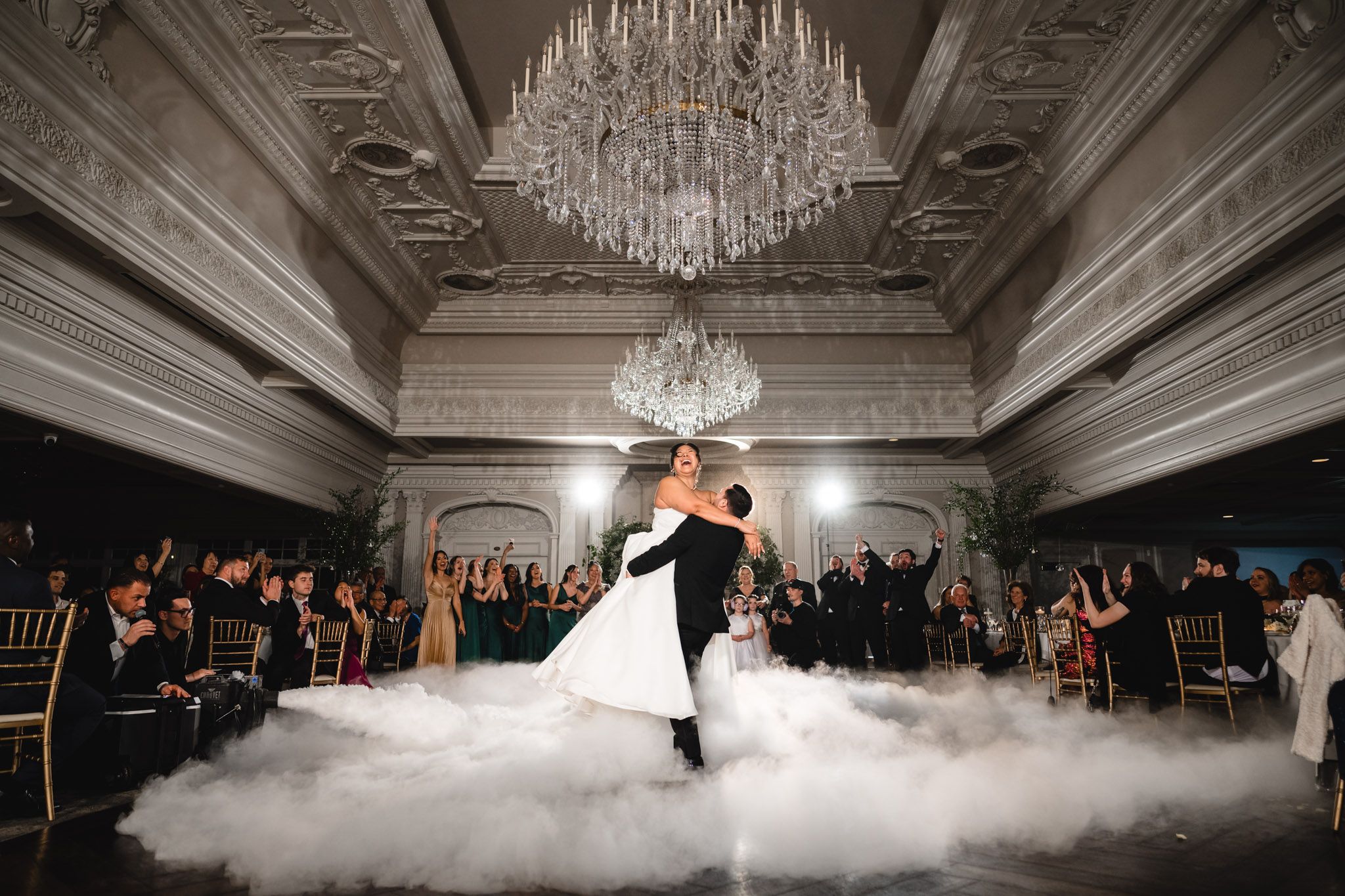 Couple performs first dance surrounded by fog effects under crystal chandelier in luxurious venue.