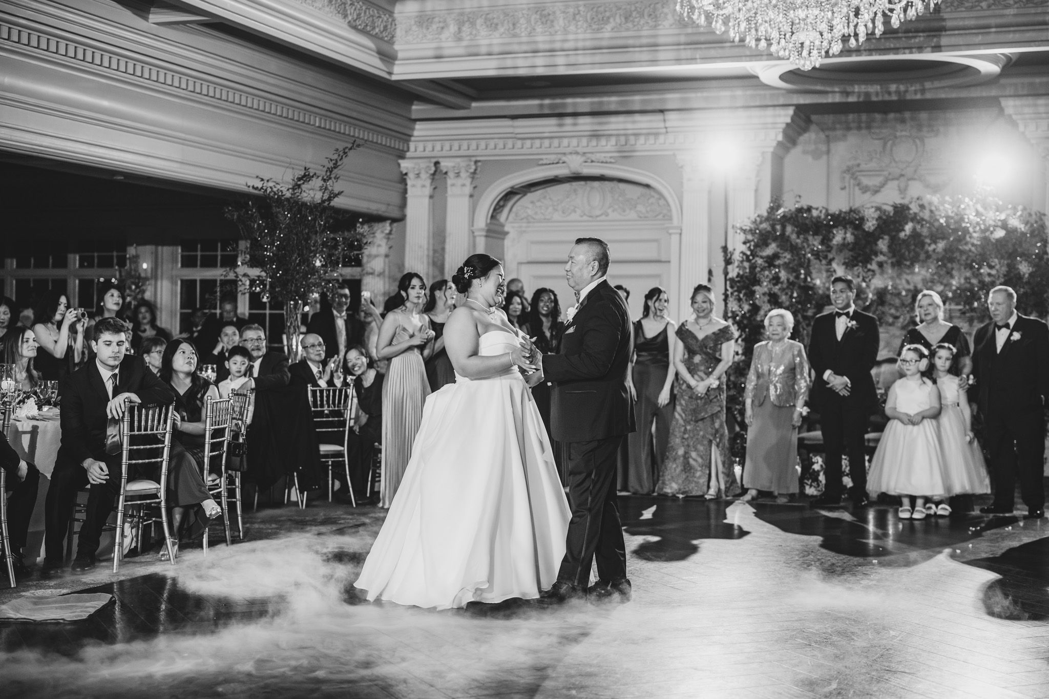 Black and white wedding reception scene in classic ballroom with guests watching first dance.