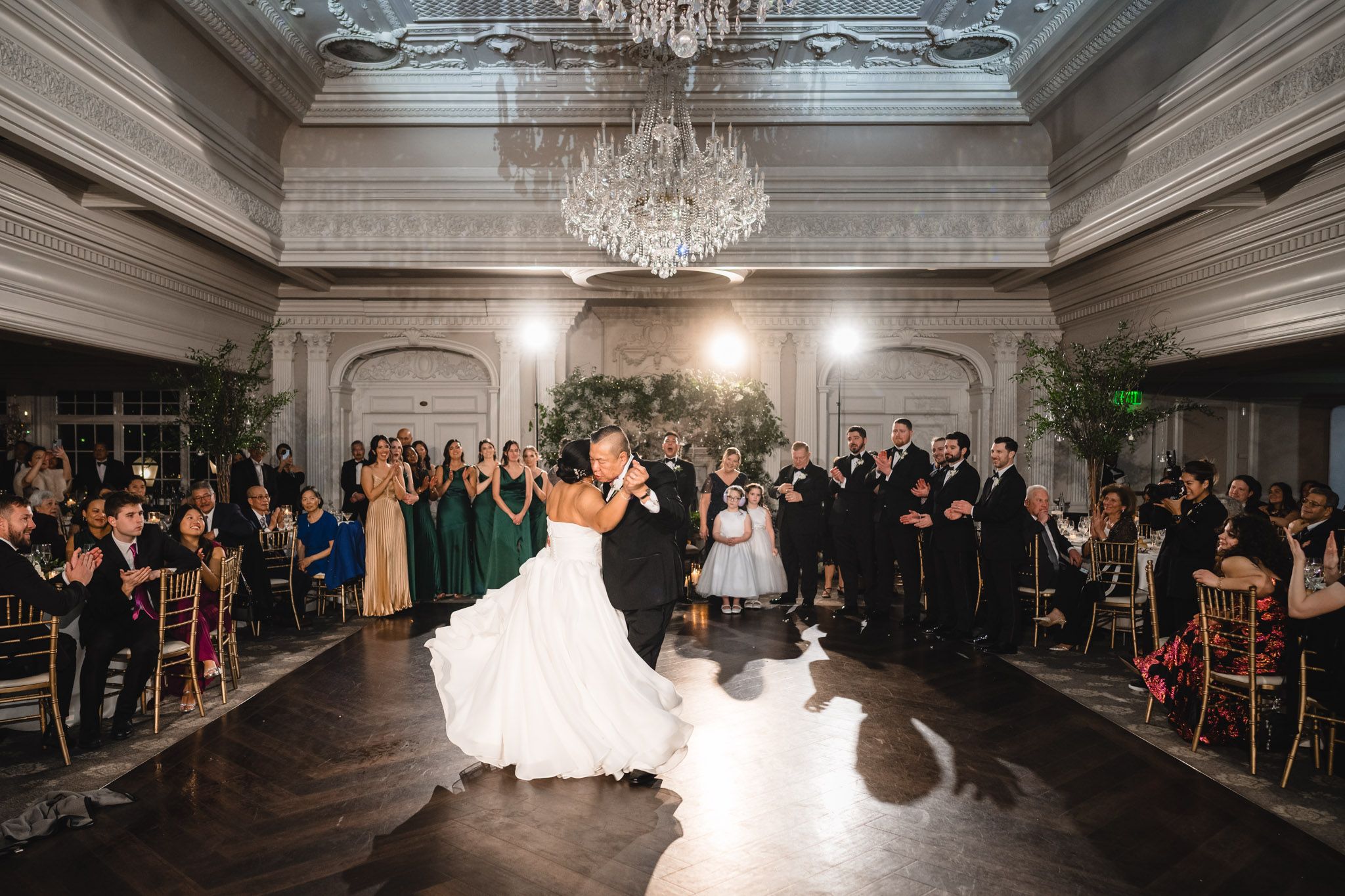 Couple shares their first dance in an elegant ballroom under a crystal chandelier while guests watch from their seats.
