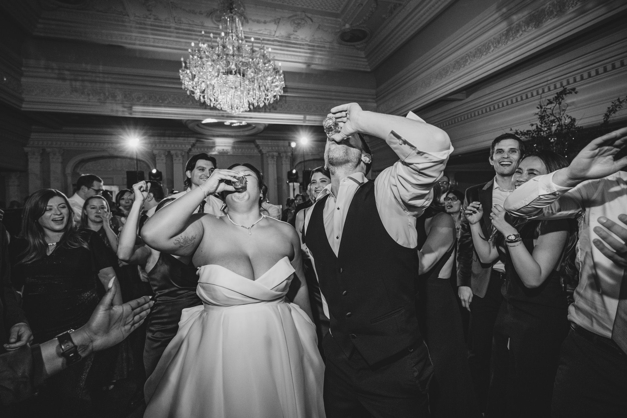 Black and white photo of wedding guests dancing and celebrating in a grand ballroom.