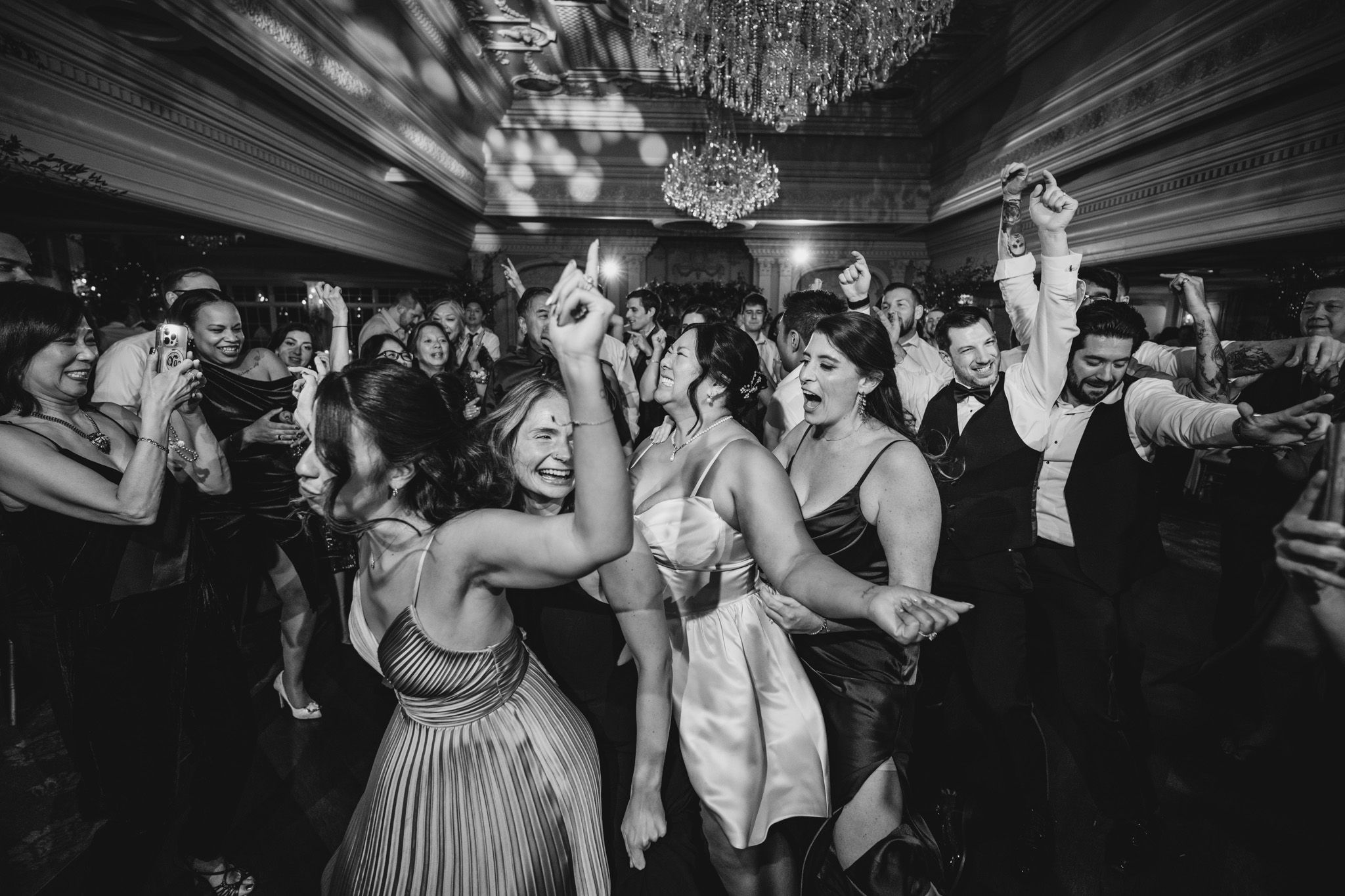 Wedding party dancing in a dimly lit ballroom under sparkling chandeliers.