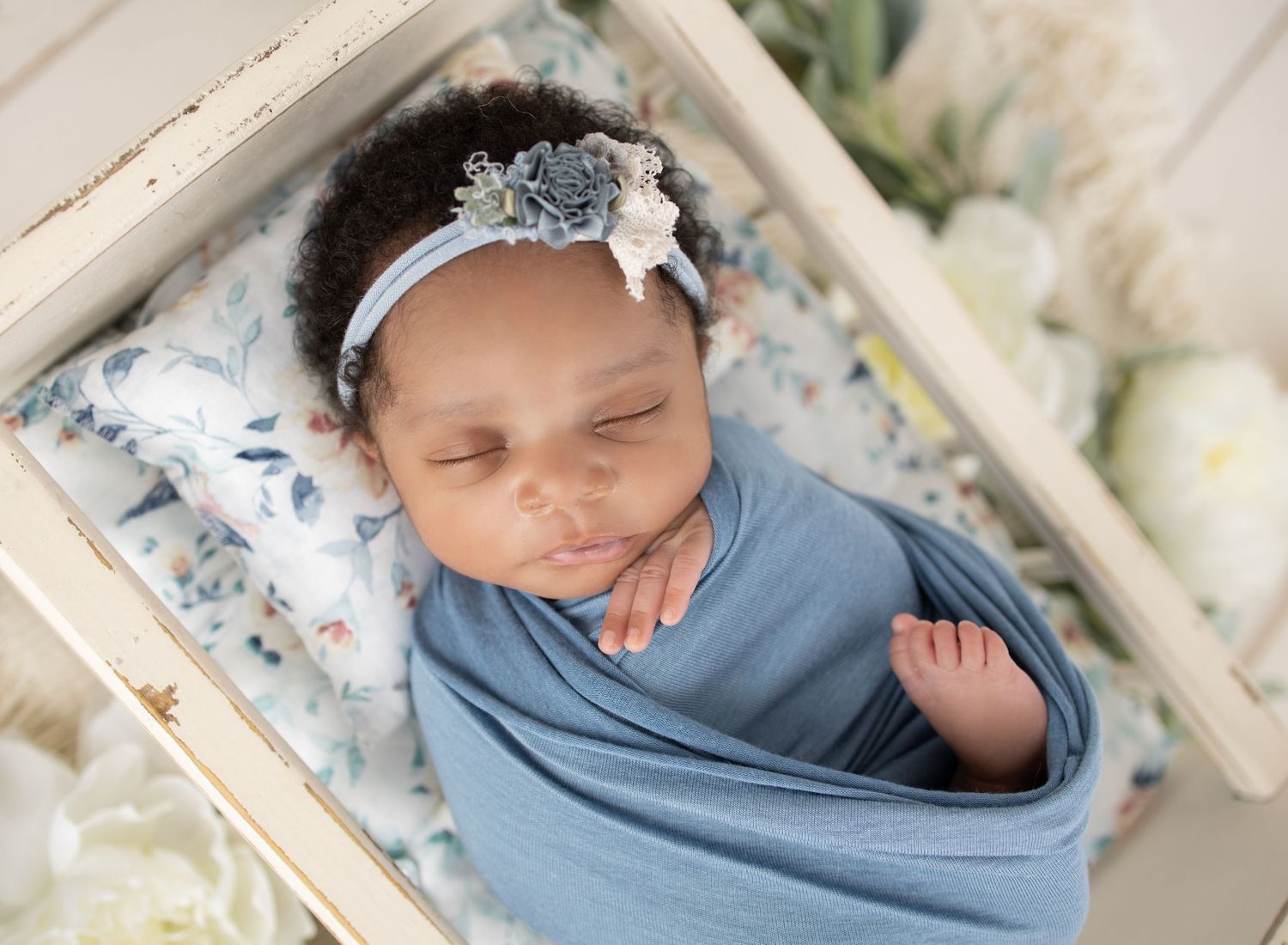 Sleeping newborn in a cozy knit sweater lies curled up in a round woven basket on white wooden floor.