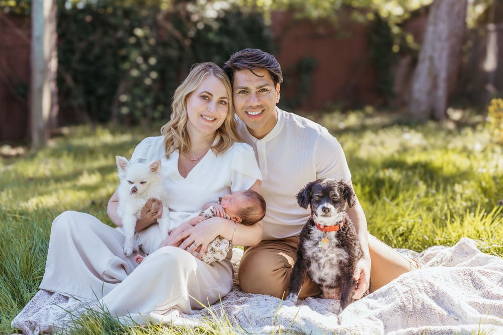 Family relaxing outdoors on blanket with newborn baby and small dog in sunny garden setting.
