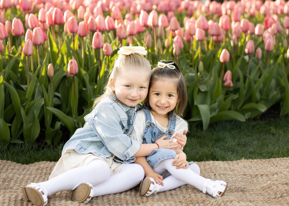 Two young children sitting together among pink tulip garden wearing spring outfits.