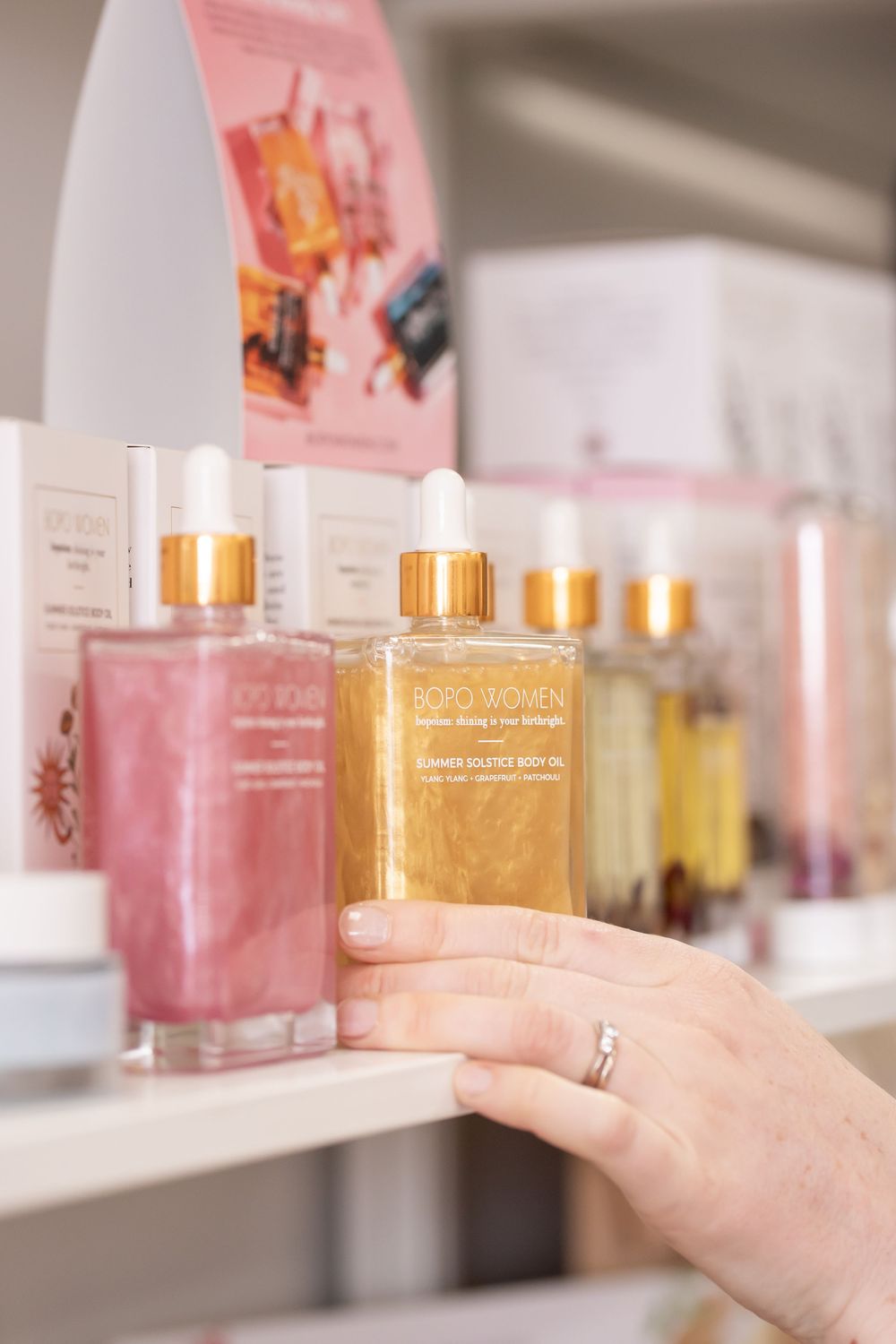 Hand reaching for gold-capped pink and amber glass bottles of beauty products arranged on white shelf.