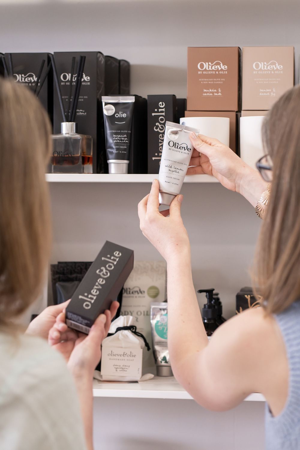 Hands reaching for skincare products on bathroom shelf displaying various Olica beauty products and packaging.
