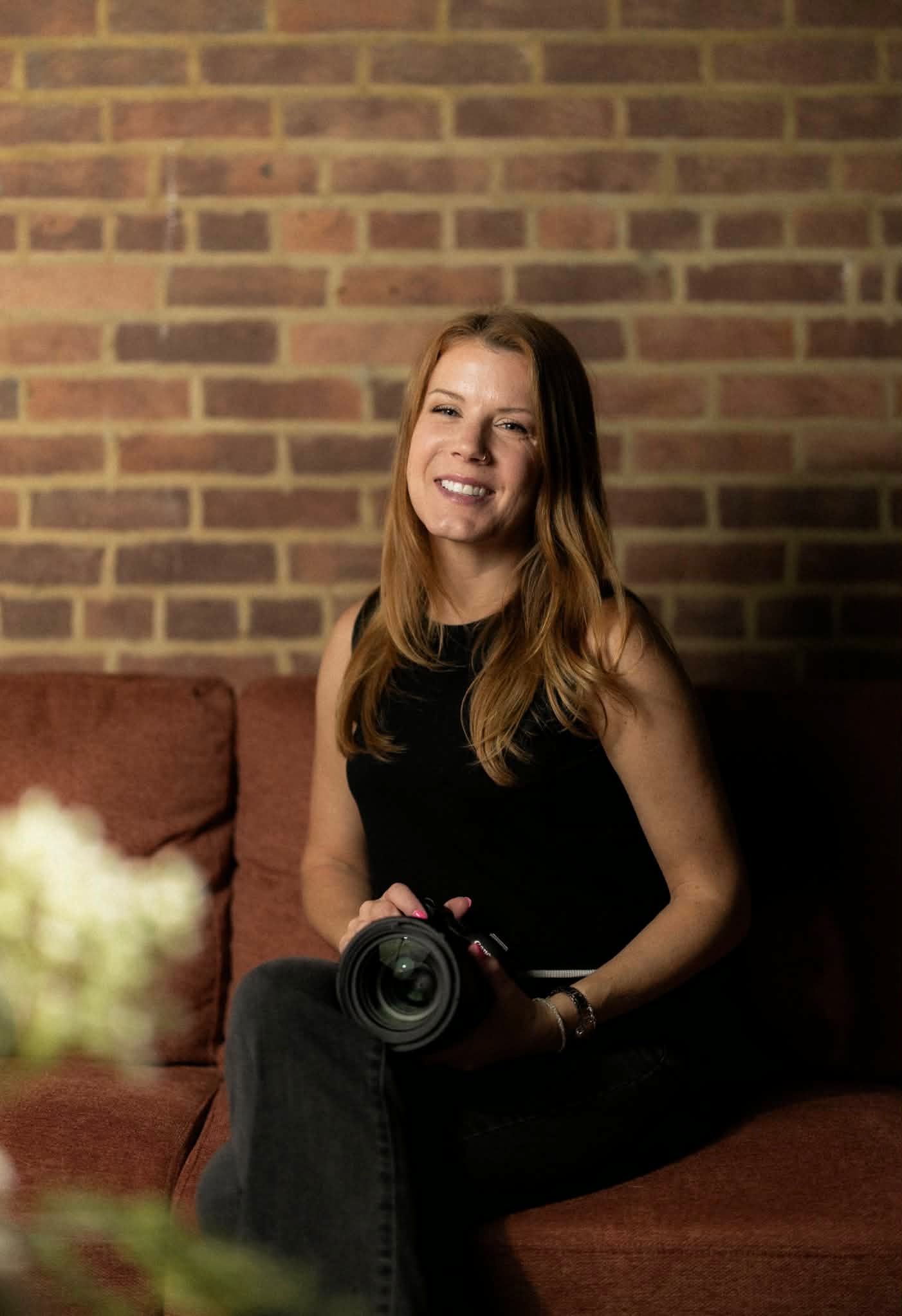 Smiling female photographer holding a DSLR camera, seated on a red couch against a brick wall backdrop.