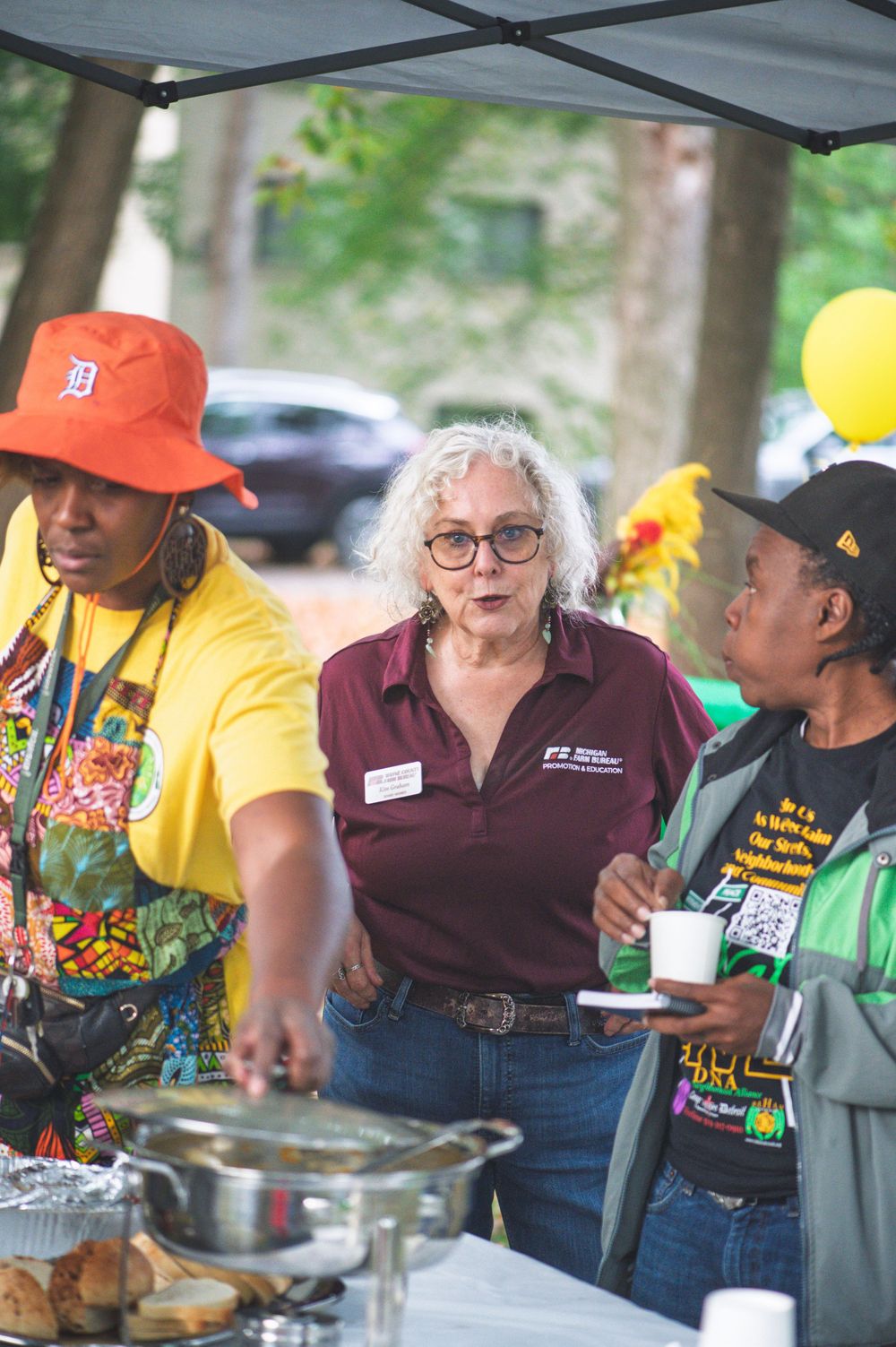 A community gathering with people serving food under a tent with colorful balloons and festive atmosphere.