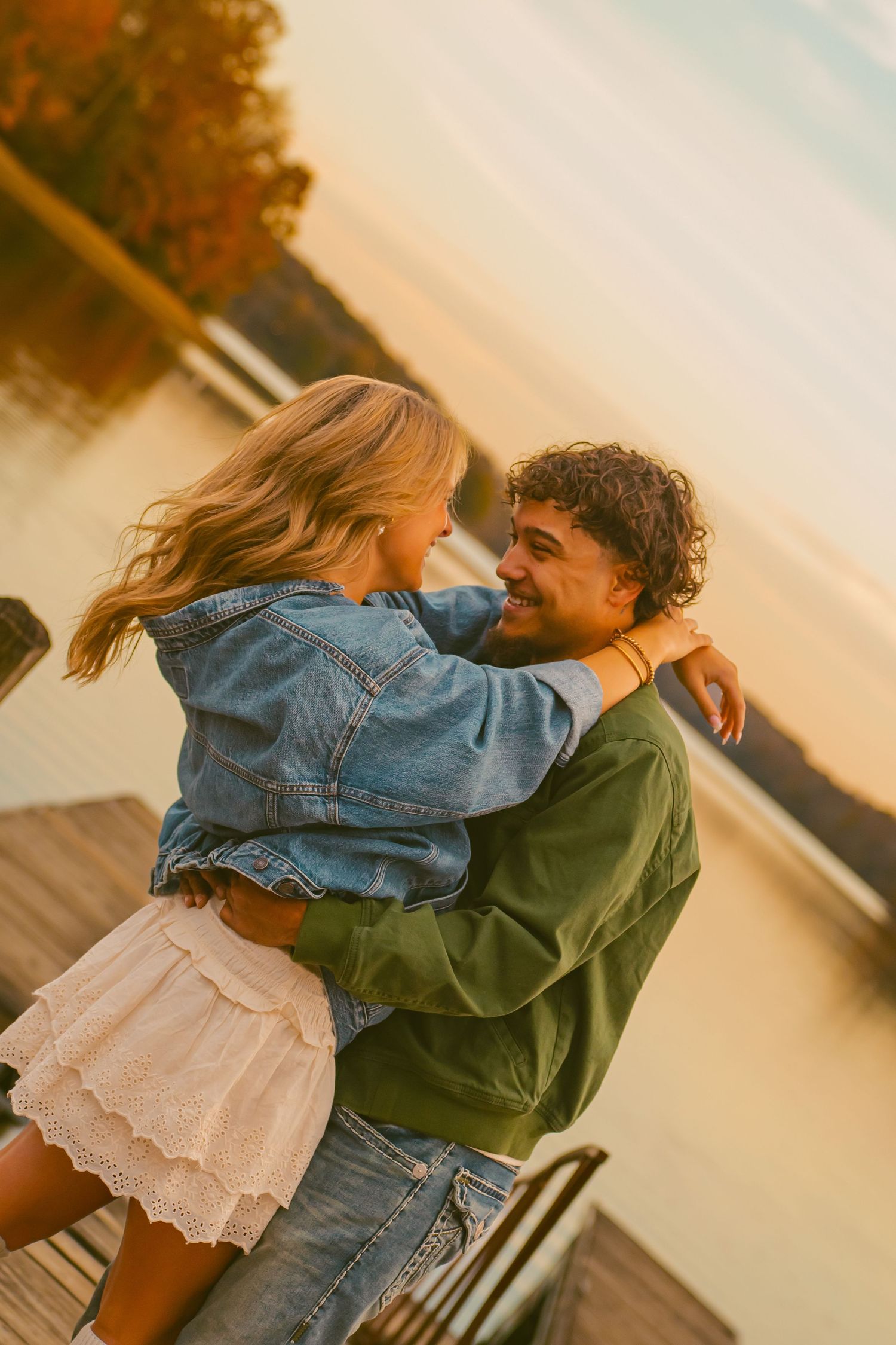 A romantic couple embraces on a wooden dock at sunset, with her in a denim jacket and white skirt, him in green.