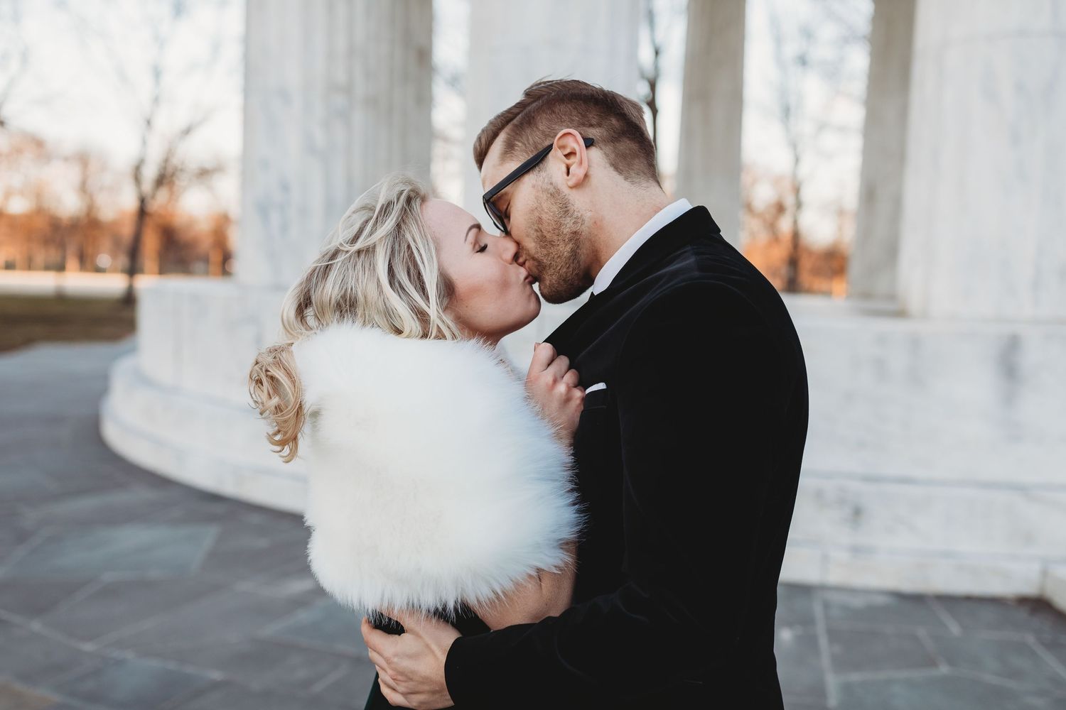 Couple sharing a kiss outdoors in winter, bride wearing white fur wrap and groom in black suit.