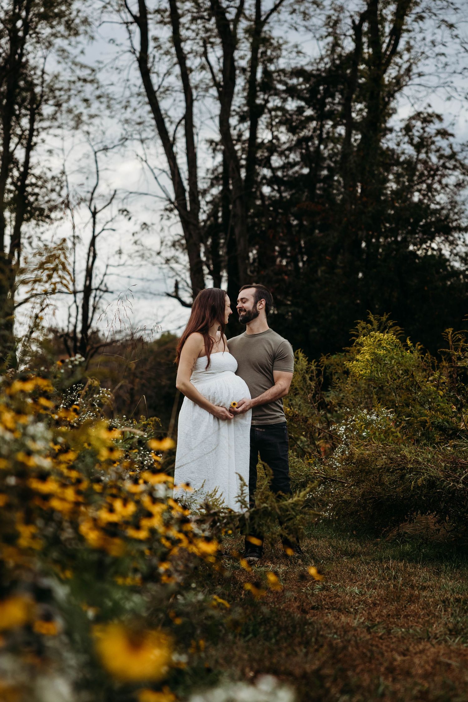 Pregnant couple standing together in autumn garden surrounded by yellow wildflowers.