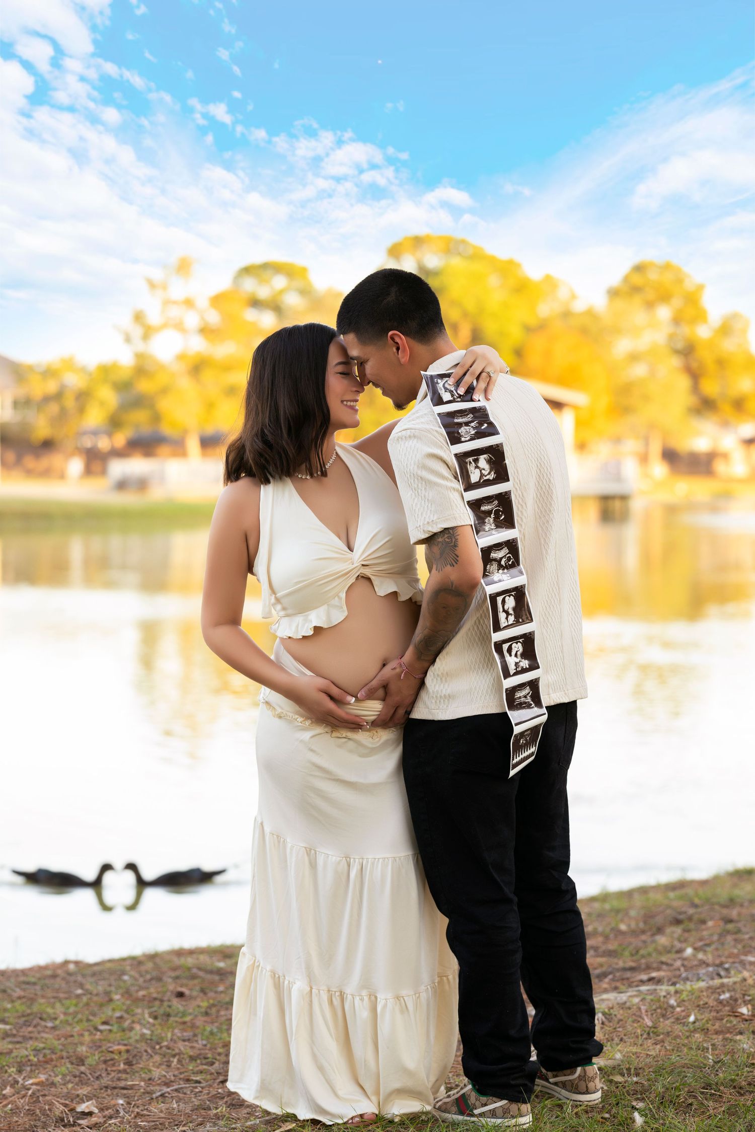 Romantic maternity photoshoot by lake with couple holding ultrasound photos.