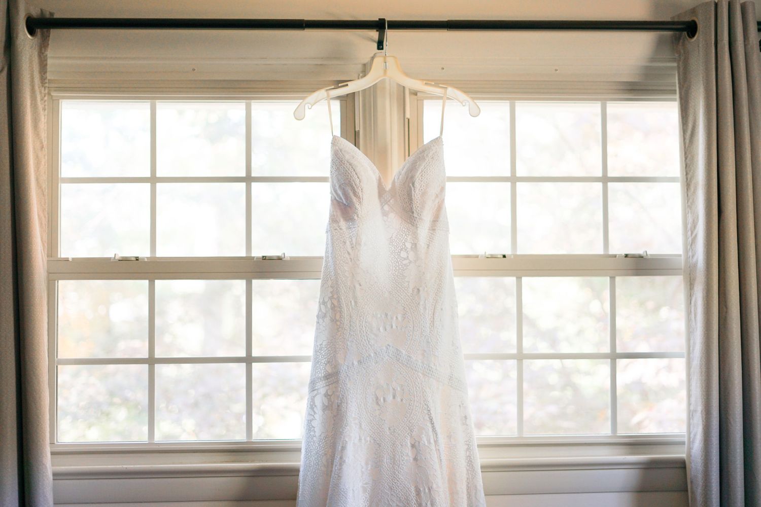 White lace wedding dress hanging in front of a sunny window.