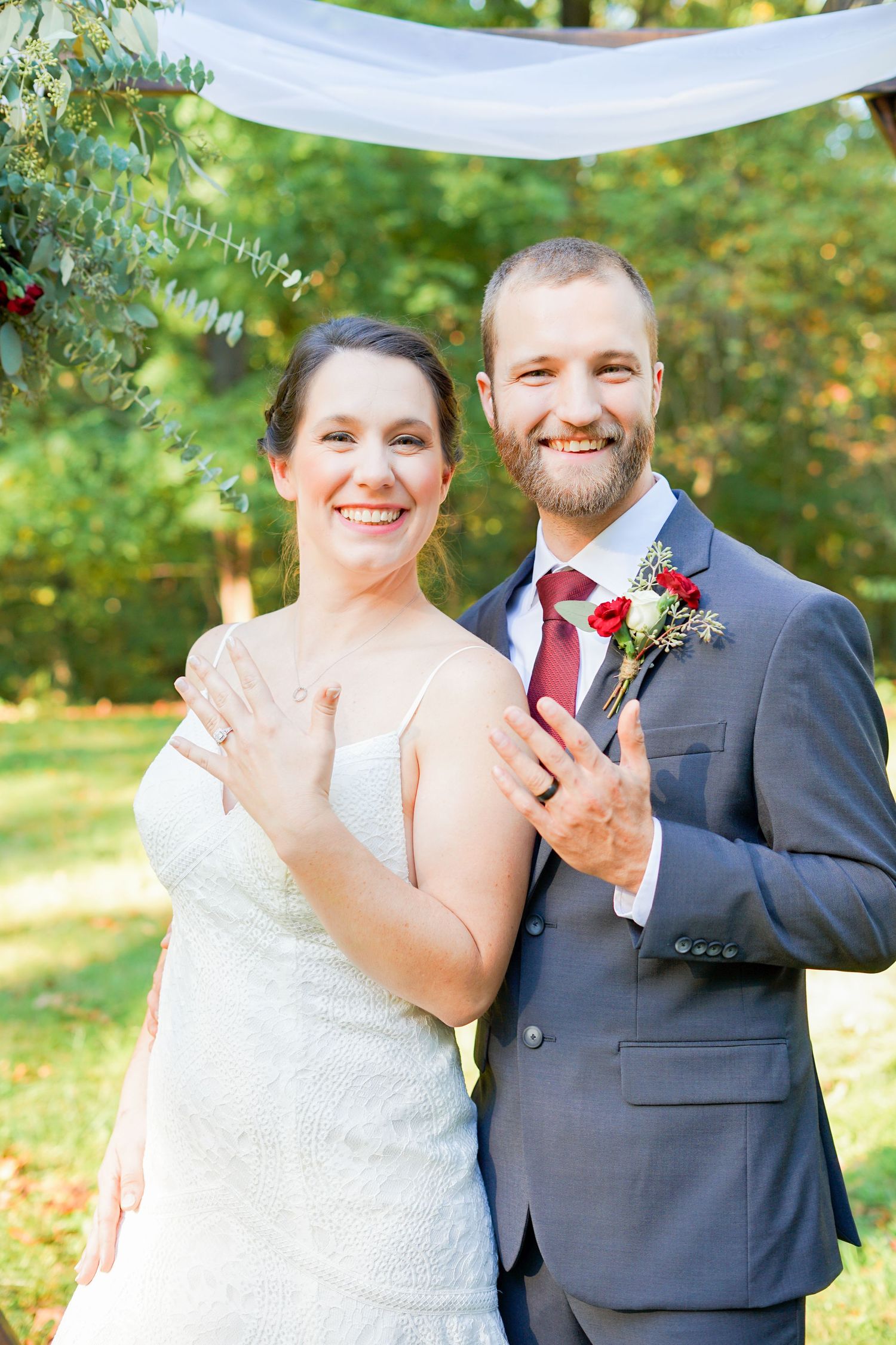 A wedding couple poses under a white drape with greenery and floral decorations outdoors.