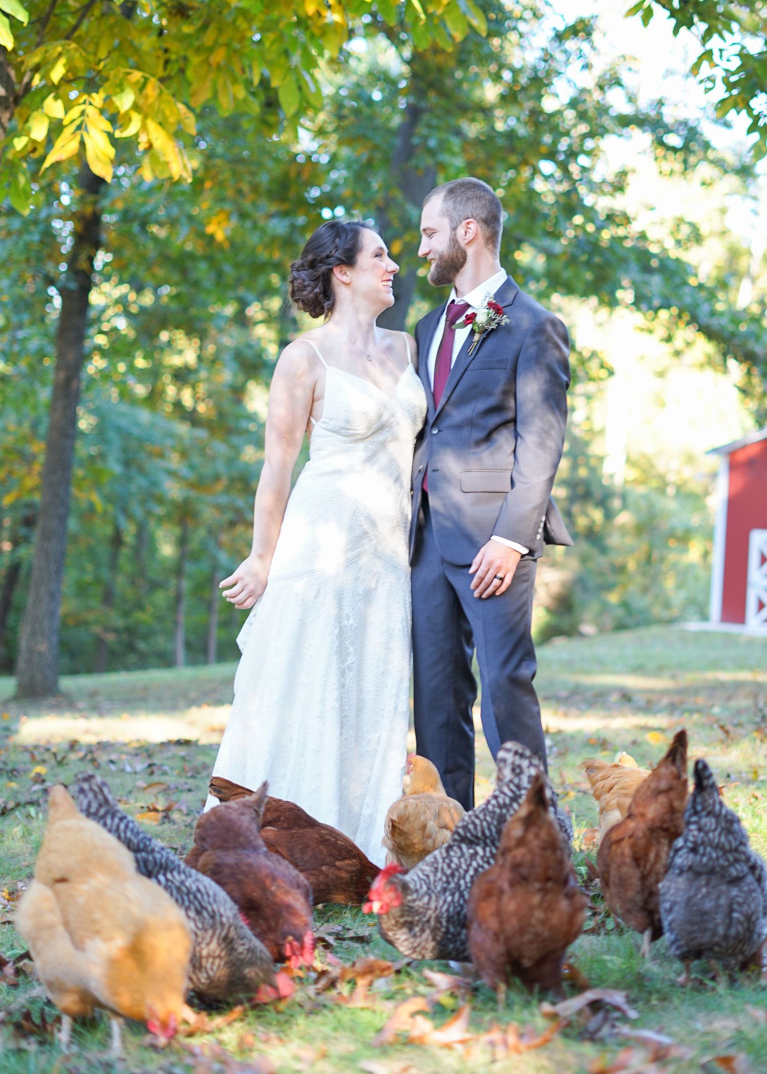 A couple in wedding attire stands outdoors surrounded by brown and red chickens in a garden setting.