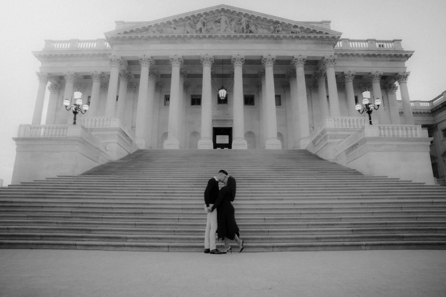 A couple stands on the steps of the U.S. Capitol building in Washington DC in a black and white photograph.
