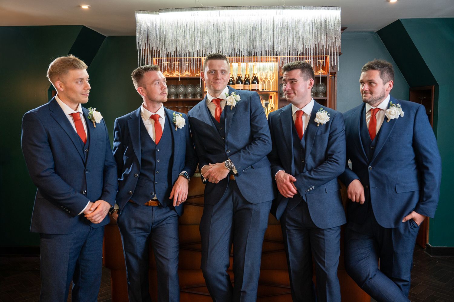 Groomsmen in navy suits and orange ties share a candid laughing moment in the 1920's bar at Chapelhouse wedding venue.