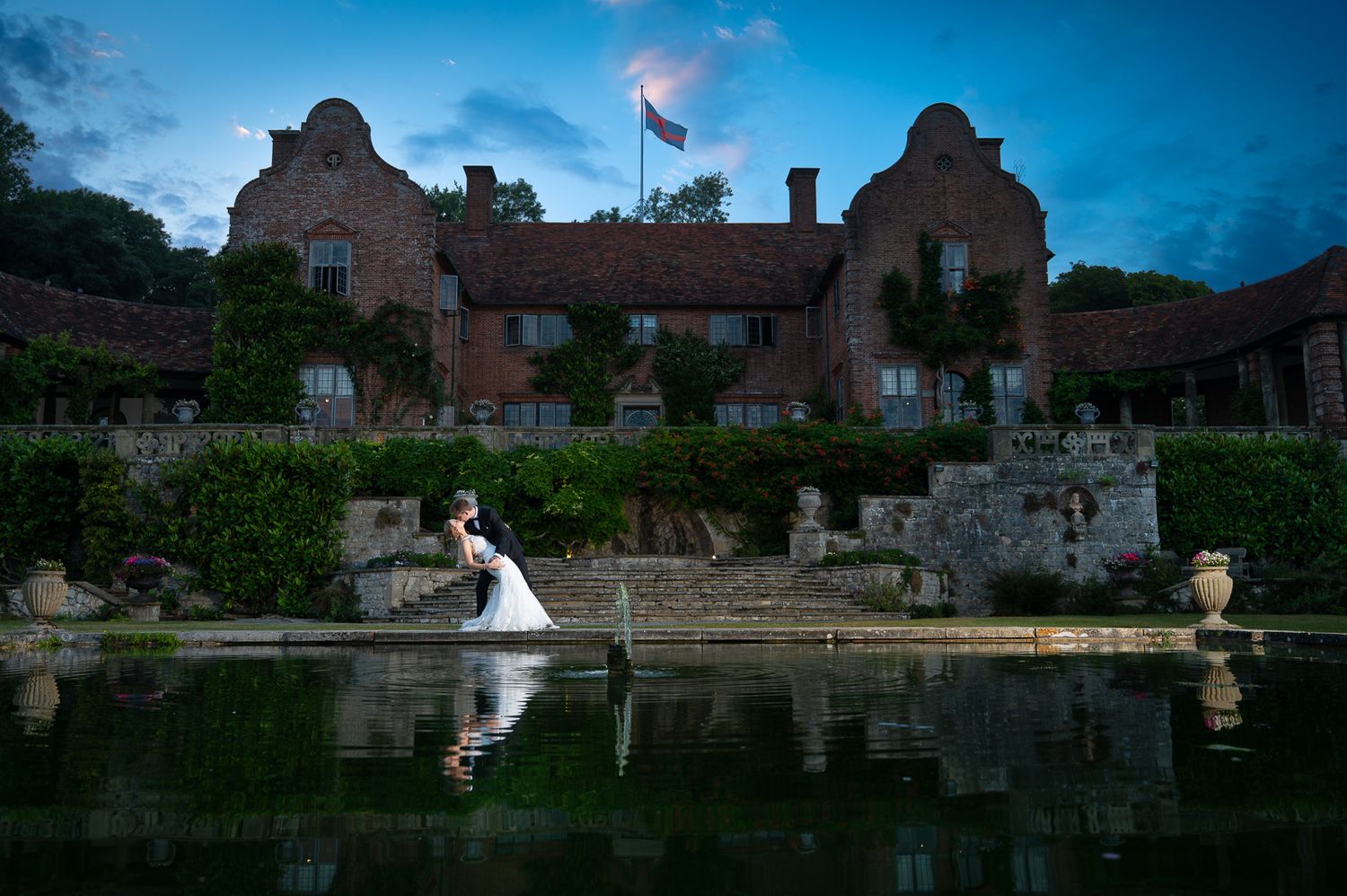 A couple in a Hollywood dip on the far side of a pond at Port Lympne, reflected under soft evening blue hour light in front of the mansion.