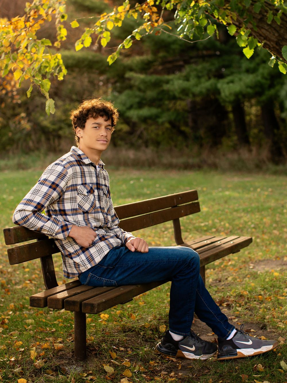 A person in a plaid shirt and jeans sits on a wooden park bench surrounded by autumn foliage.