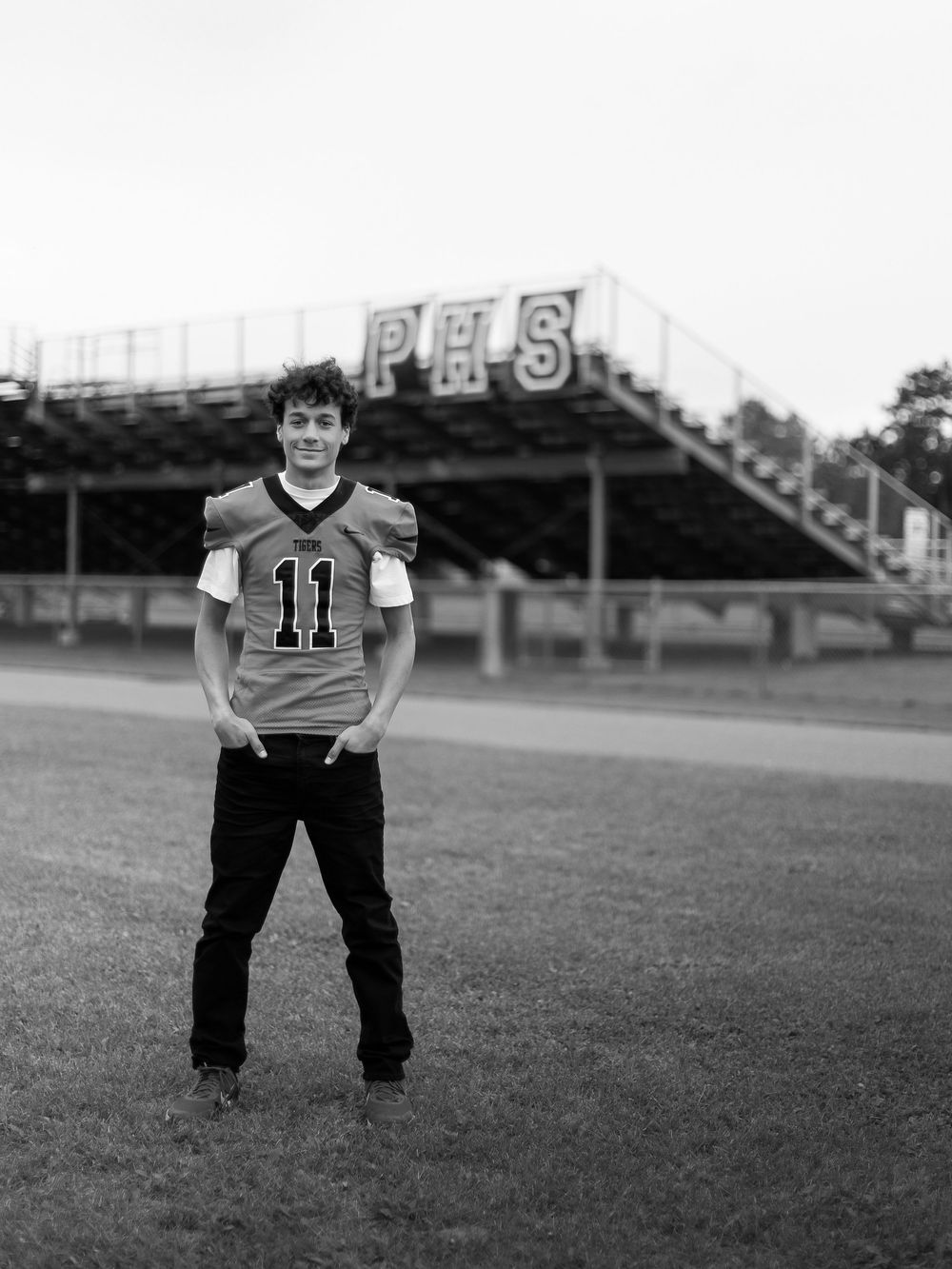 Black and white photo of a football player wearing jersey number 11 standing on a field with stadium bleachers in background.