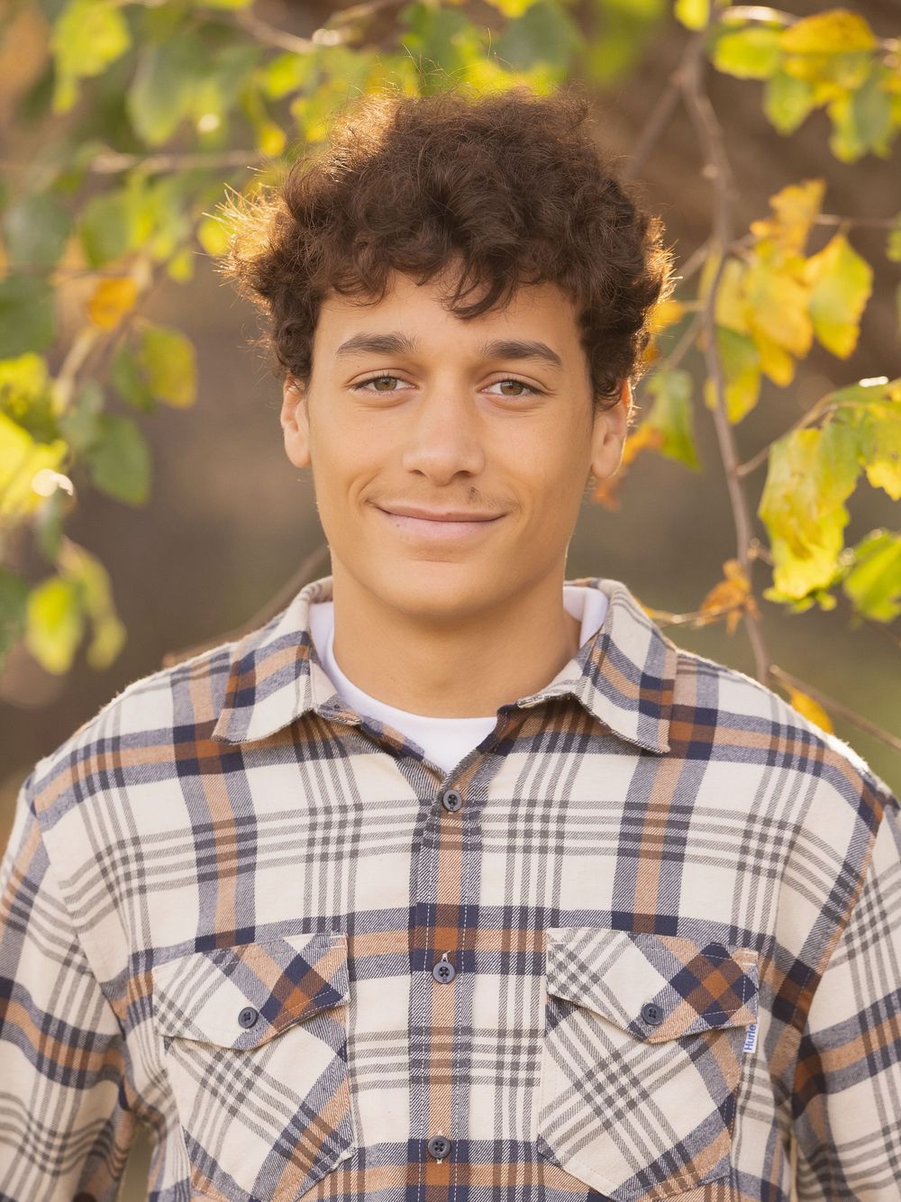 A person in a plaid flannel shirt poses outdoors with autumn leaves in the background.