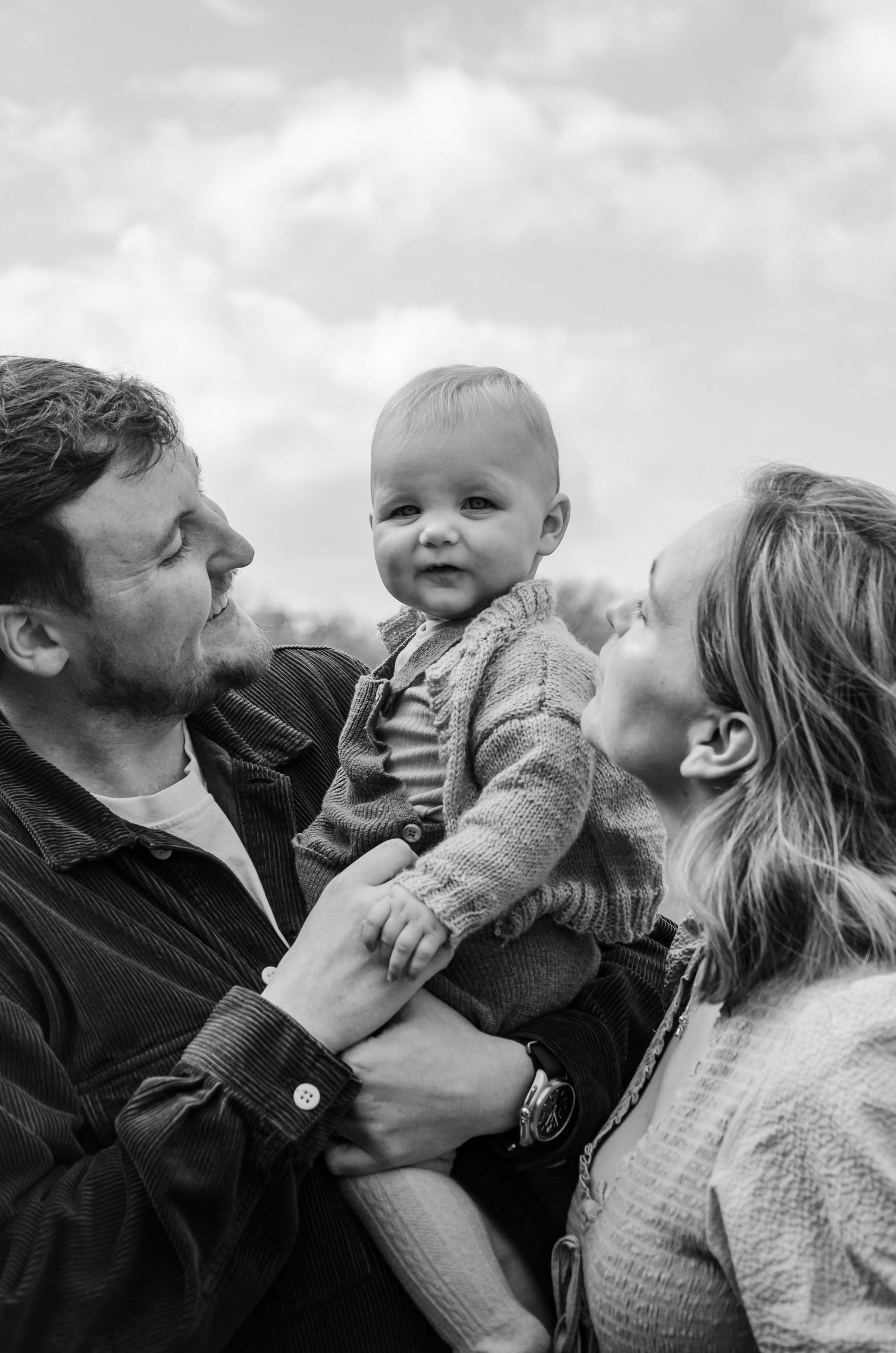 Black and white candid family moment with baby outdoors under cloudy sky.