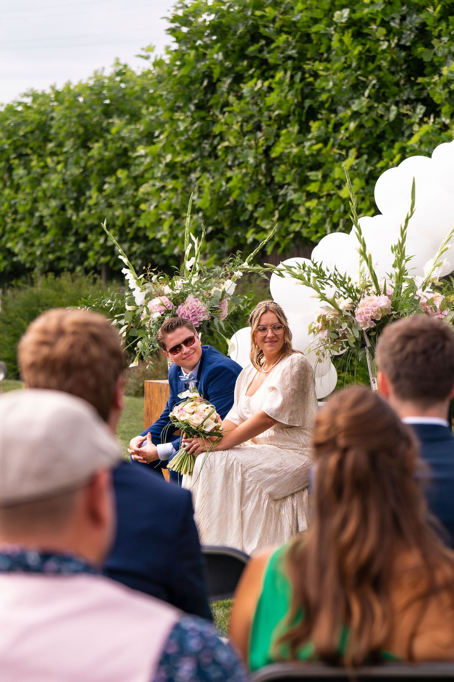 Outdoor wedding ceremony with white balloon arch and lush green hedge backdrop.