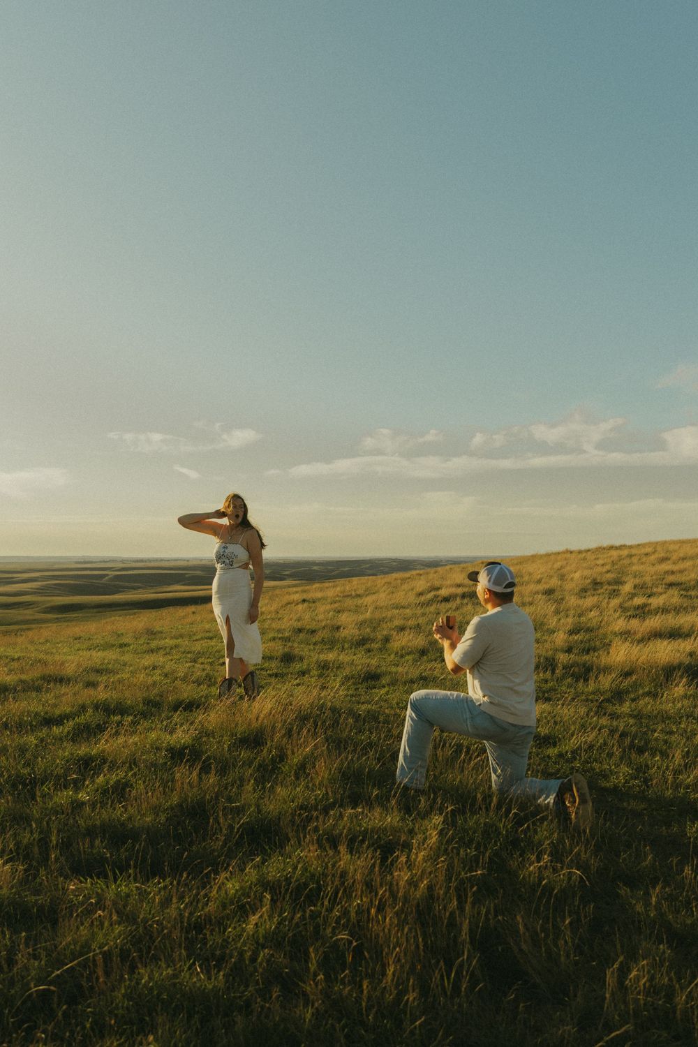 Surprise proposal moment captured during sunset on grassy hilltop.
