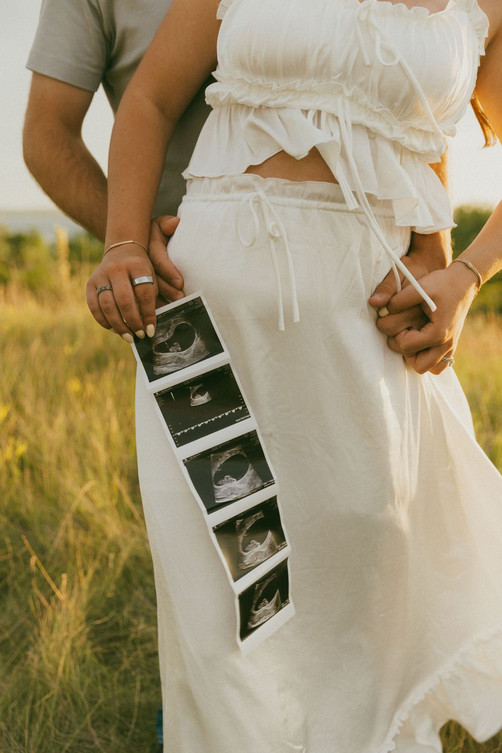 Someone in a white dress holds ultrasound photos in a sunlit meadow.