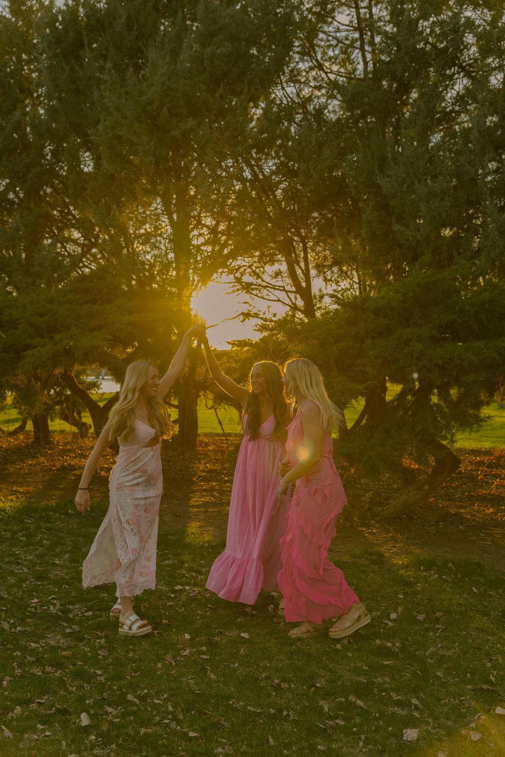 Three friends in flowing dresses dance together in a golden sunset beneath towering trees.