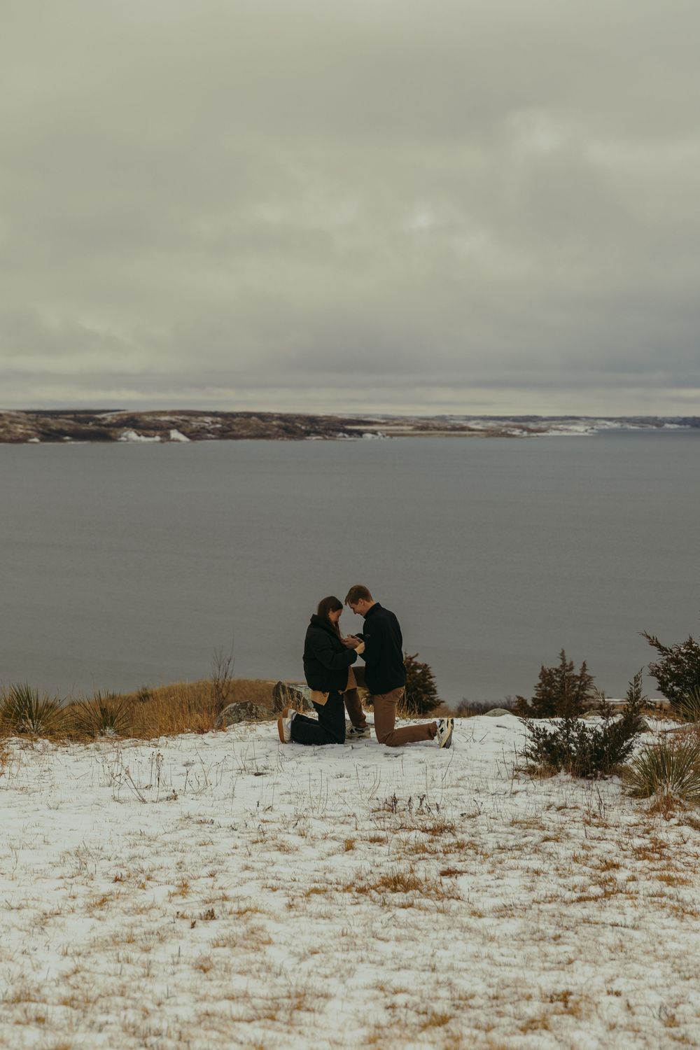 Couple sharing a marriage proposal moment on a snowy cliffside overlooking a vast gray lake under cloudy winter skies.