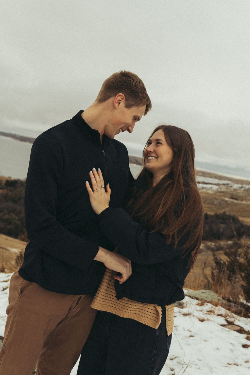 A smiling couple embraces outdoors in a snowy winter landscape, wearing cozy sweaters, gazing at each other romantically.