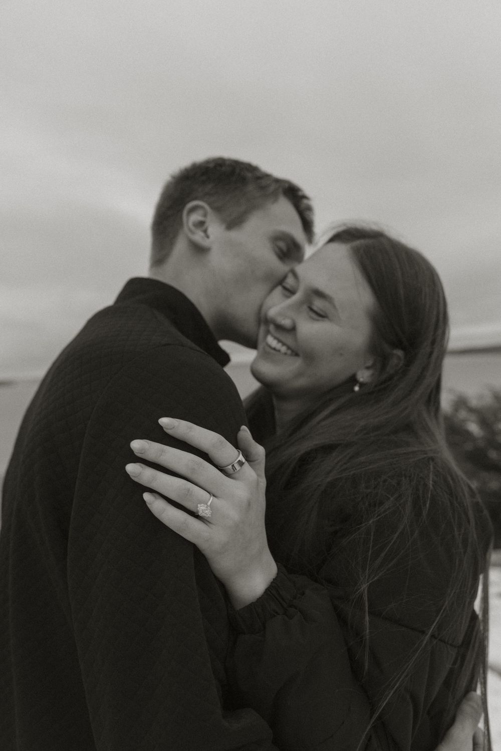 Black and white photo of a smiling couple embracing, woman wearing engagement ring, romantic outdoor portrait.