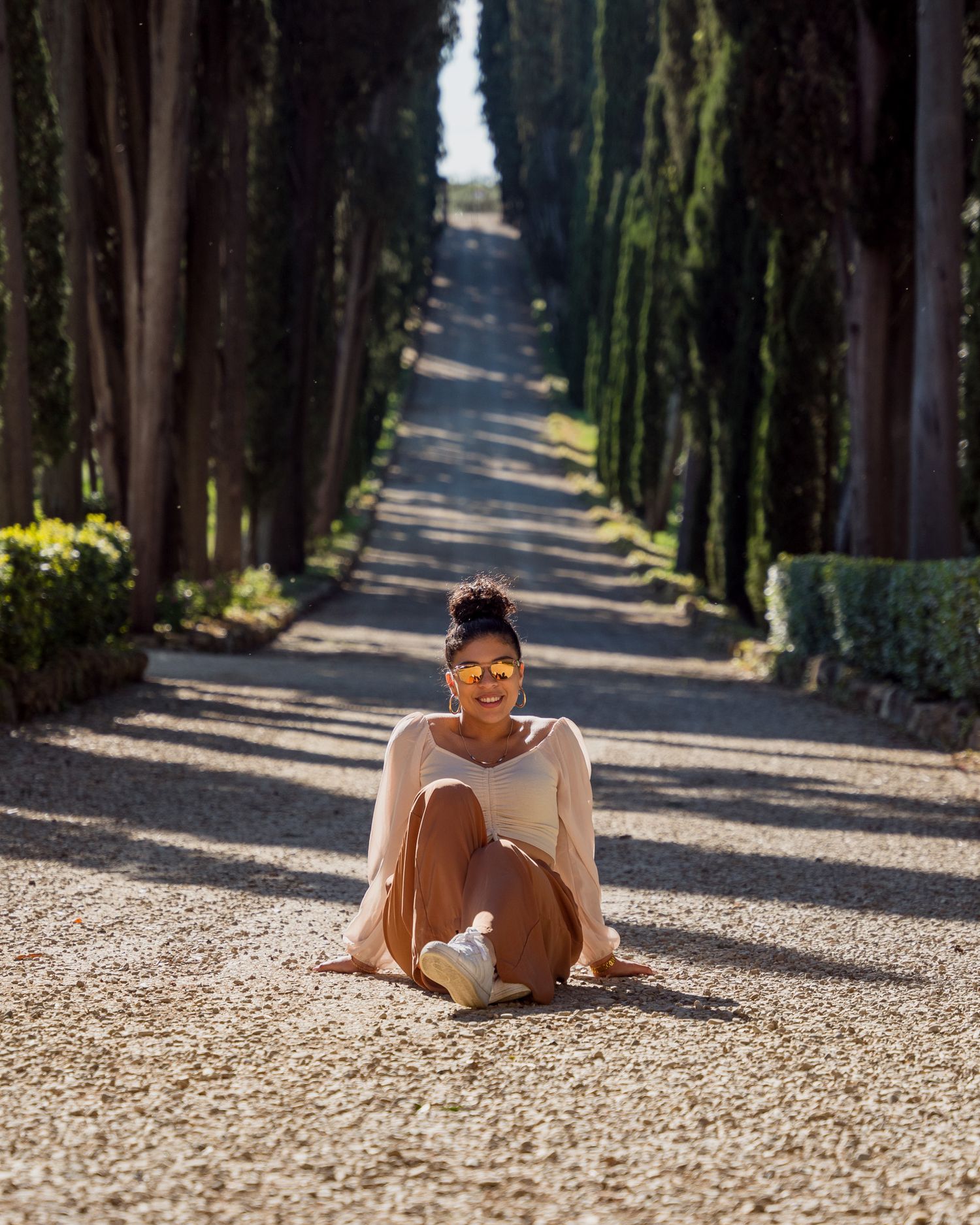 Person sitting on a gravel path lined with tall cypress trees in warm sunlight.