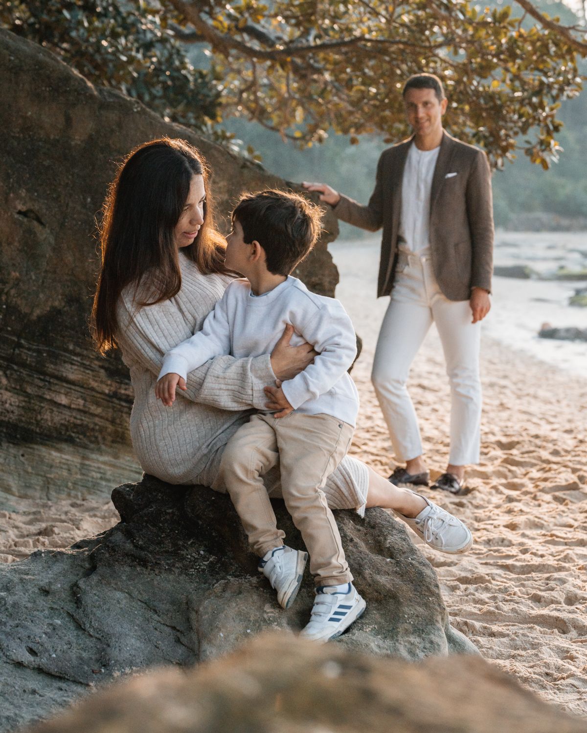 son-sitting-on-moms-lap-and-looking-at-her-while-dad-is-standing-and-smiling-behind-them-during-a-family-photoshoot-in-b