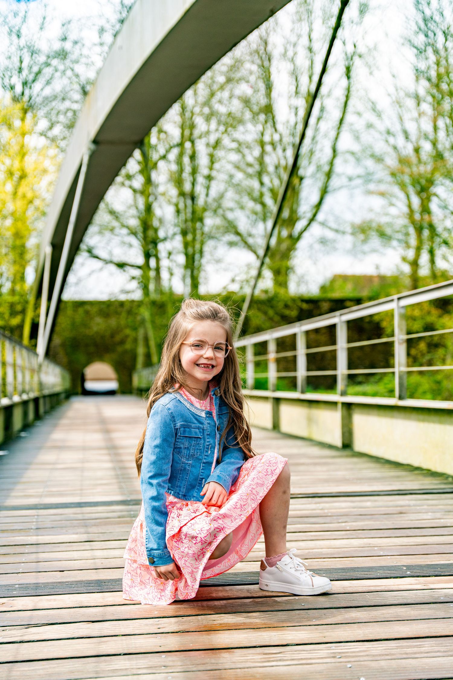 Child in blue jacket and pink dress sitting on modern wooden bridge walkway.