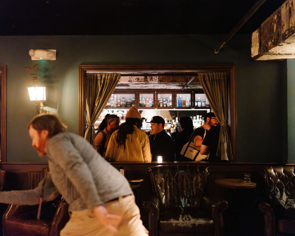A dimly lit bar interior with patrons gathered by a window while someone in a light jacket moves in the foreground.