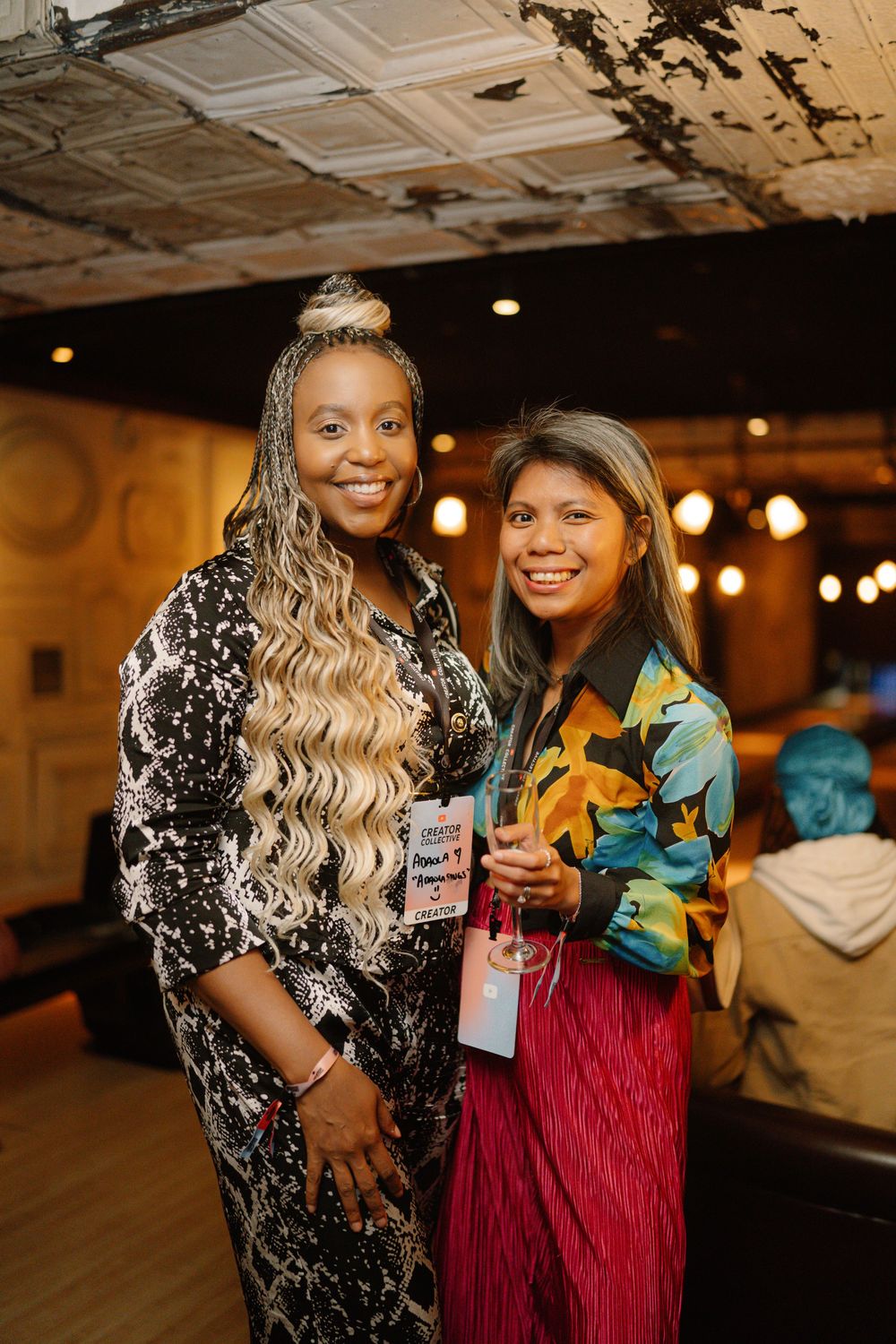 Two friends in colorful outfits pose together at an indoor event with decorative ceiling and ambient lighting.