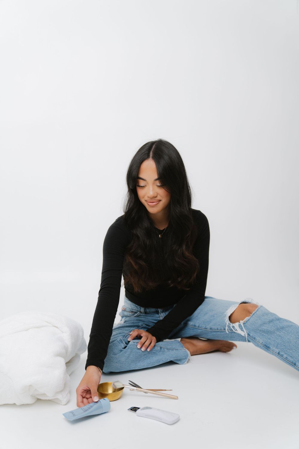 Woman in black sweater and distressed jeans sits on white floor with phone and cards beside her.