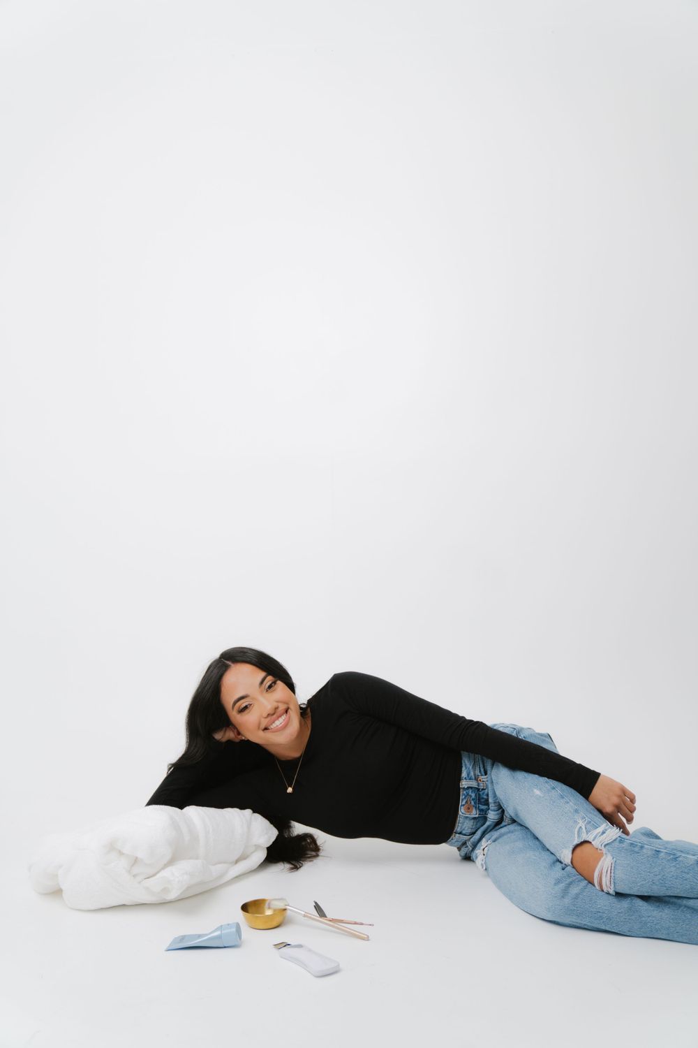 A person in a black sweater and ripped jeans relaxes on a white floor with small blue baby shoes nearby.
