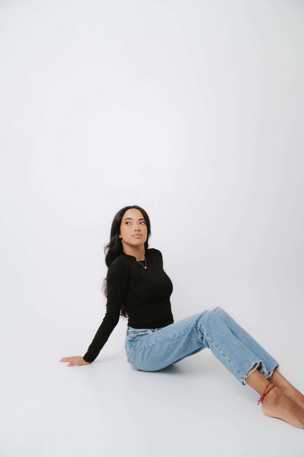 Woman in black long sleeve top and light jeans poses casually on white studio floor background.
