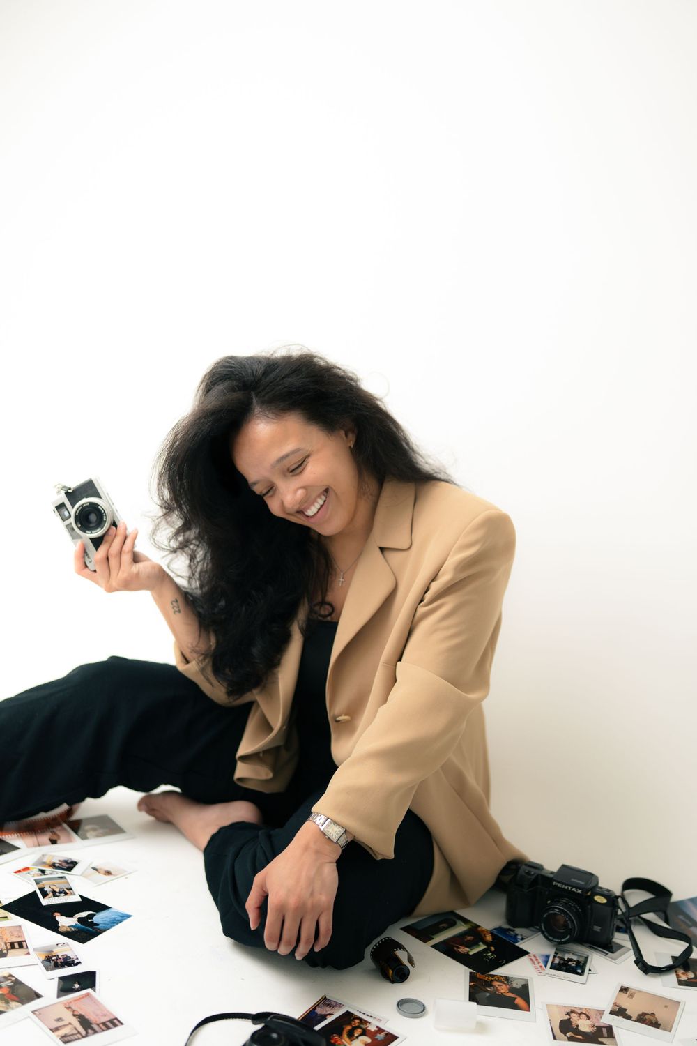 Person in beige blazer sits with vintage cameras and photographs scattered around on white background.