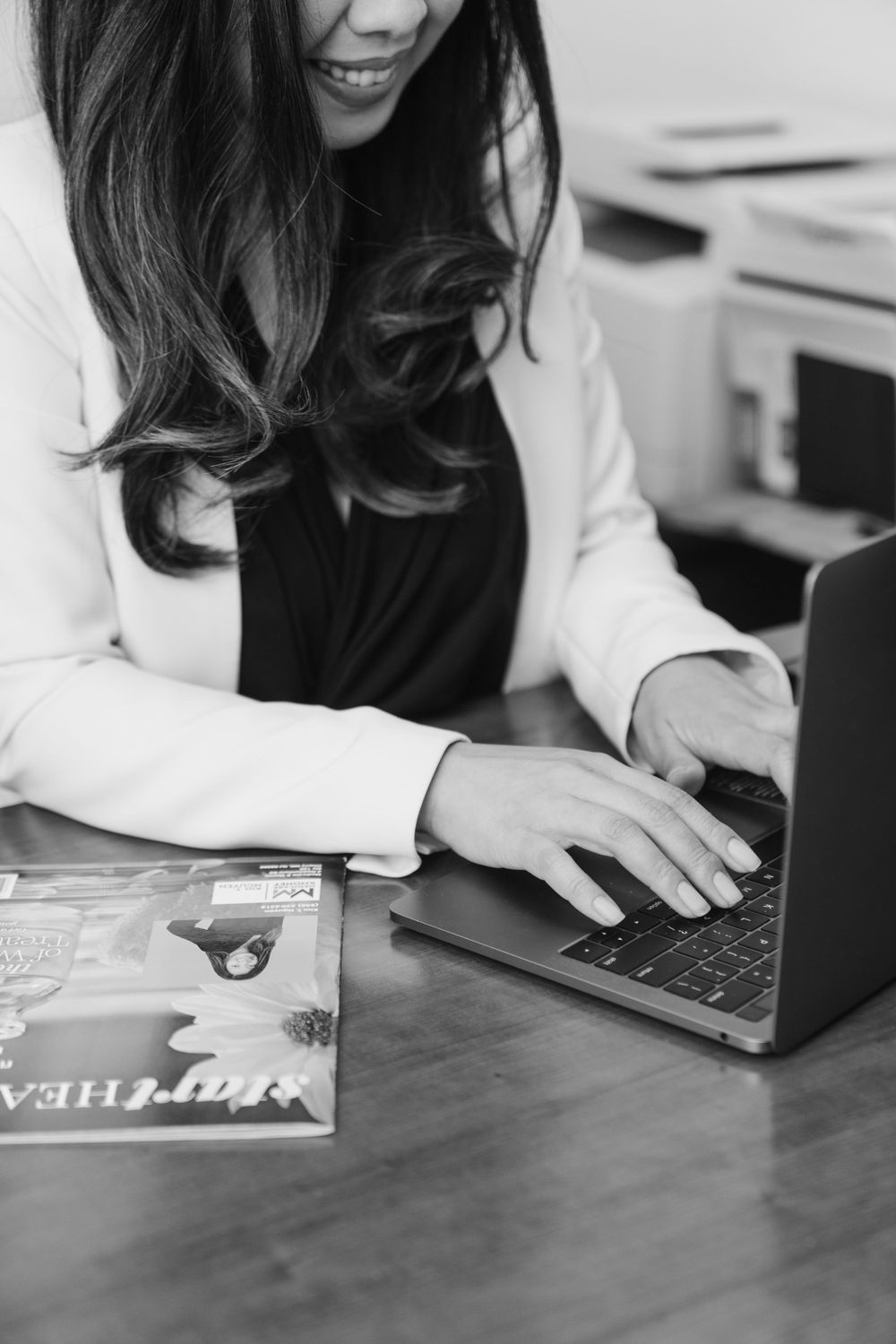 Black and white image of someone typing on laptop with book on wooden desk.