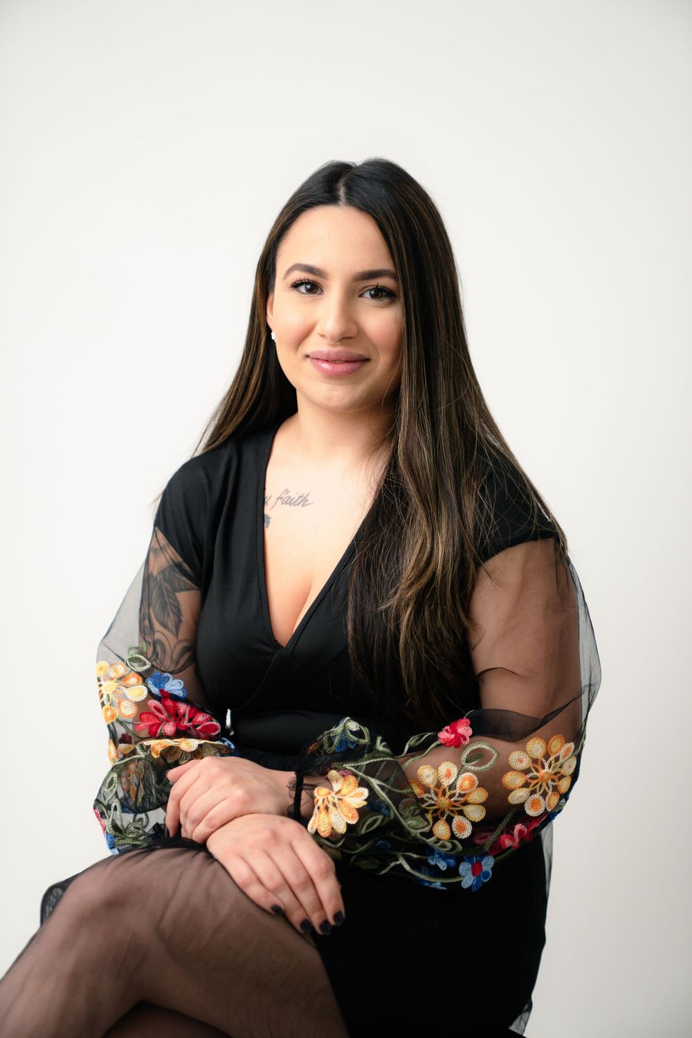 Person in black dress with sheer floral sleeves poses seated against white background for professional portrait.