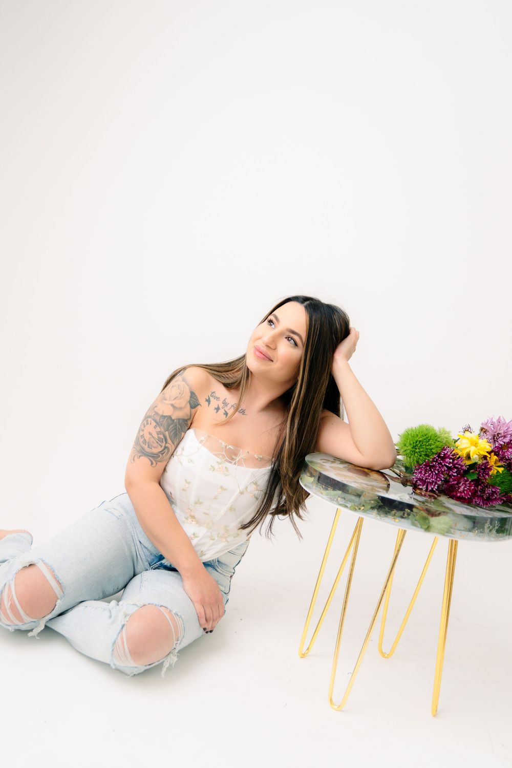 Stylish woman in ripped jeans and white top relaxes on floor next to modern side table with colorful flower arrangement.